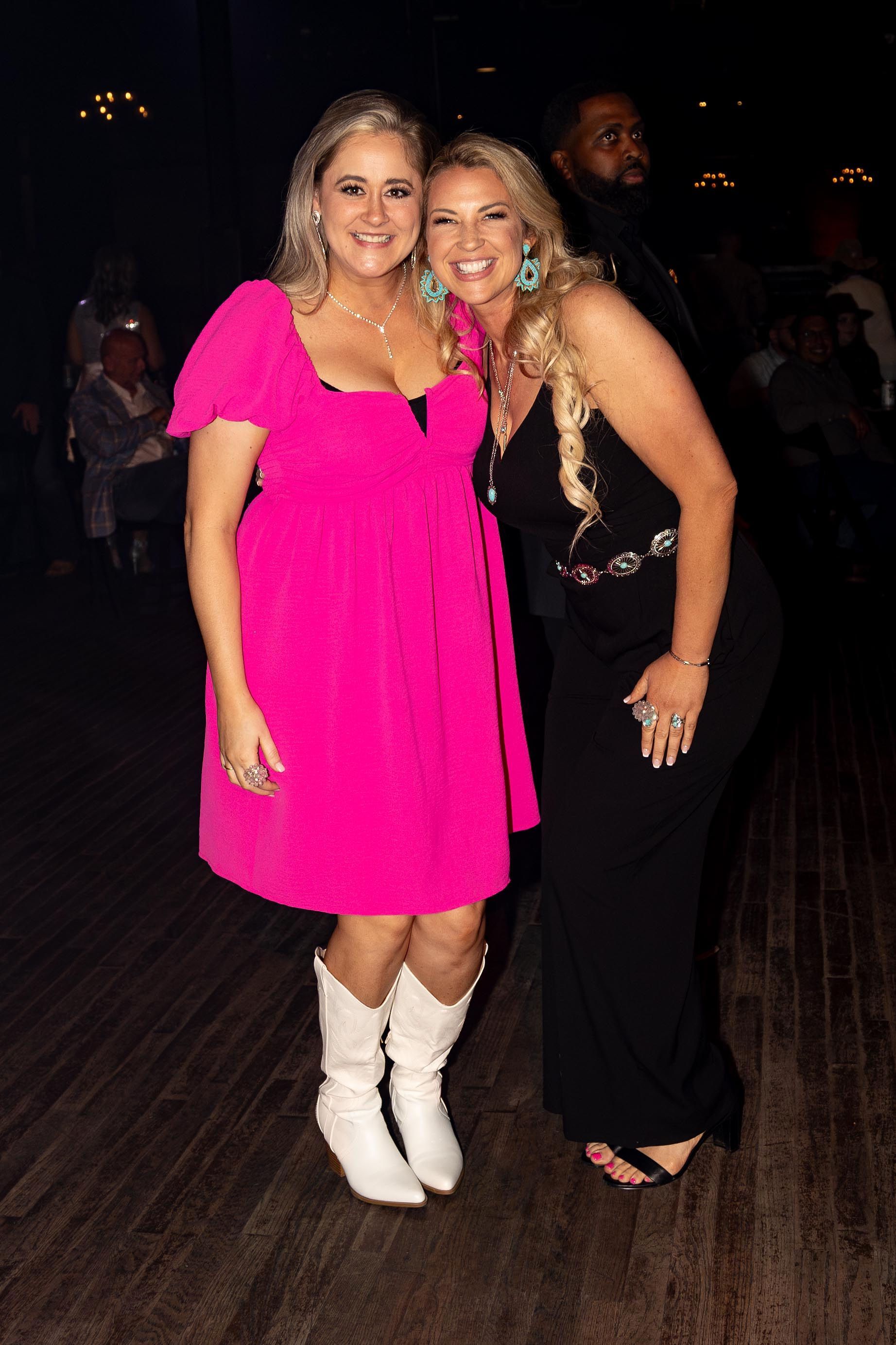 Two women smiling, one in pink dress and white boots, the other in black dress, posing on a wooden floor.