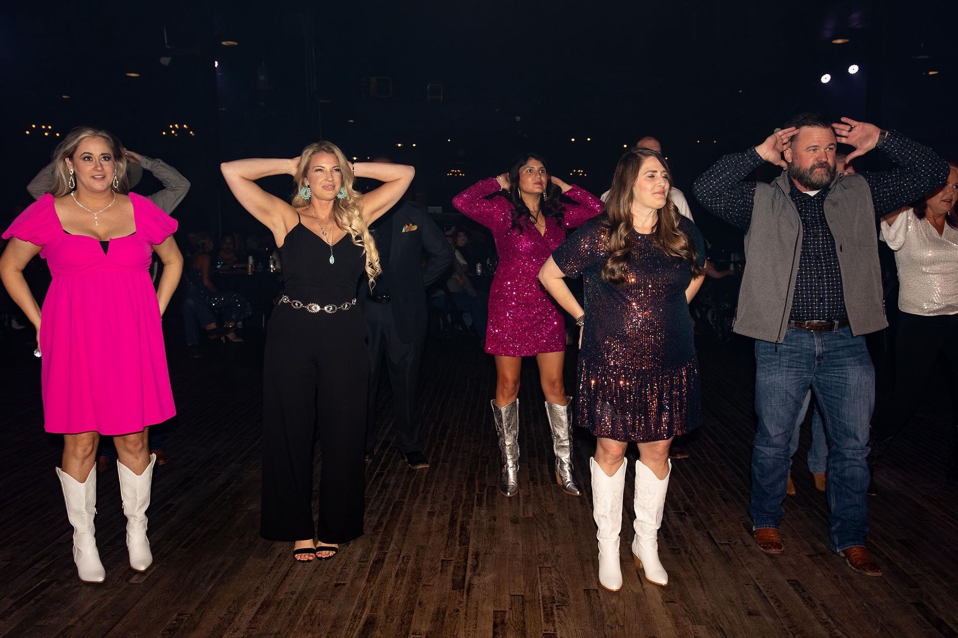 People dancing in a dark room, arms up. Women in pink, black, and sequined dresses, white boots. Man in vest, jeans.