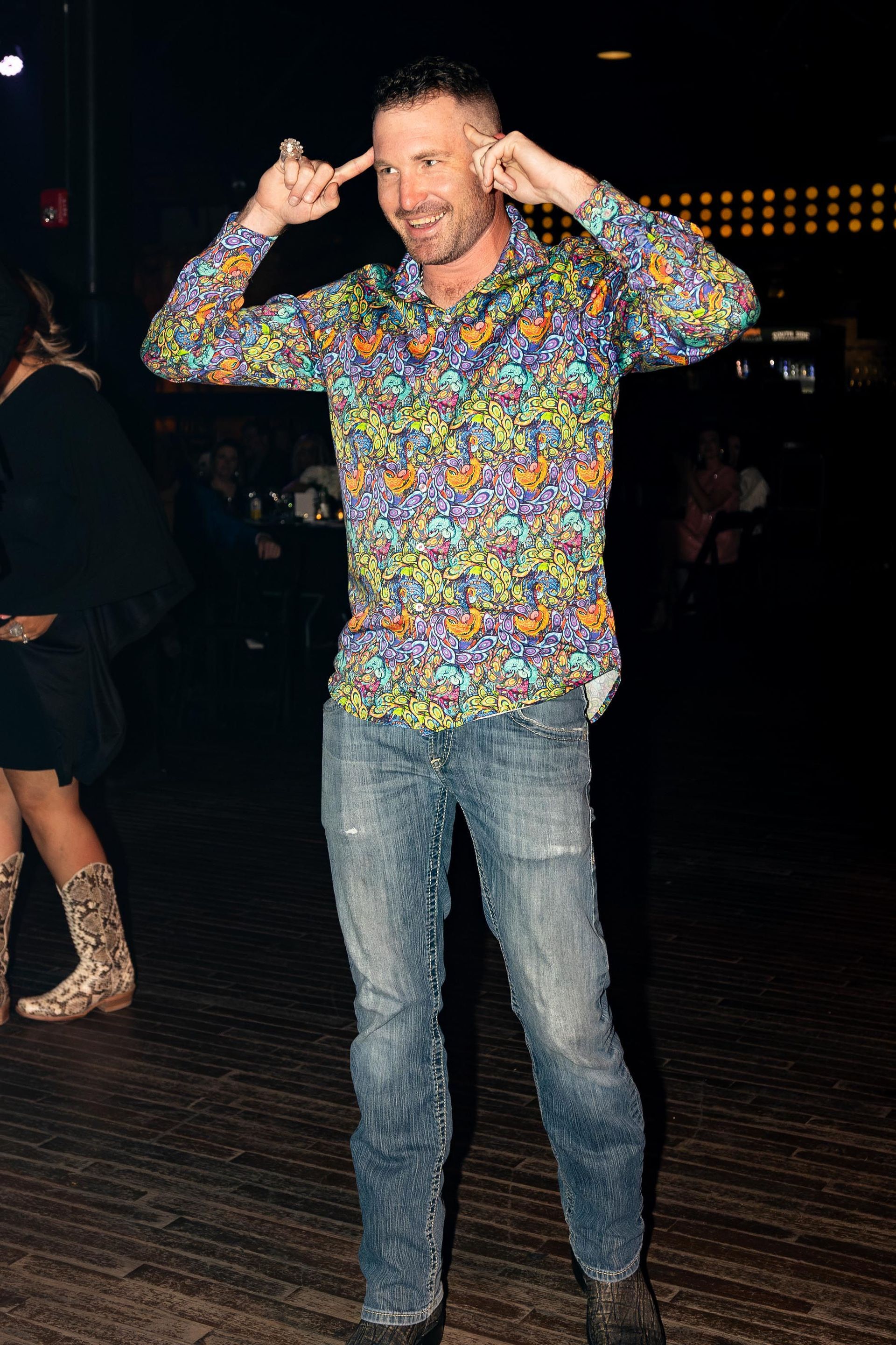 Man in colorful shirt and jeans, hands near head, making a playful gesture. Dark setting.
