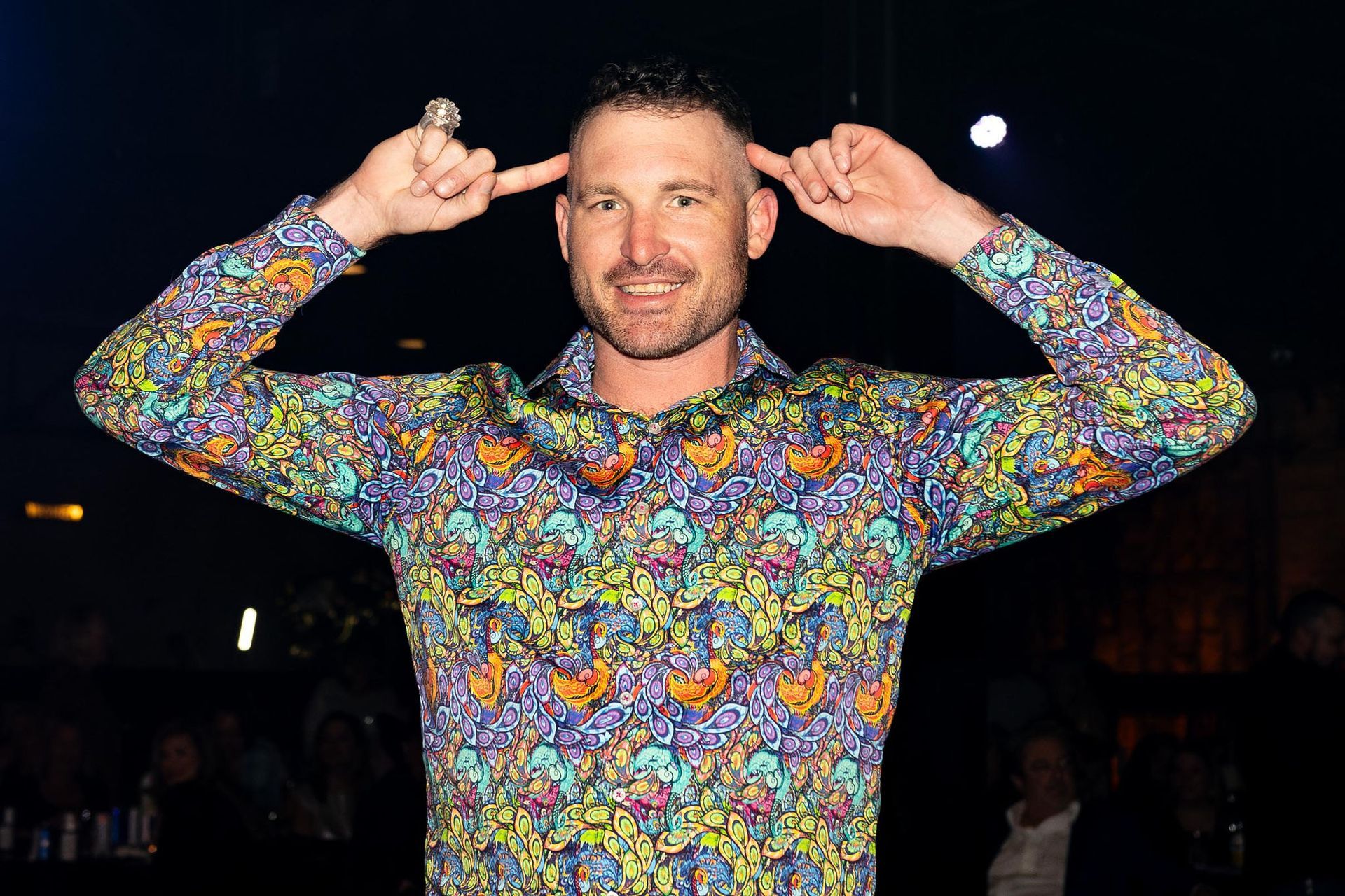 Man wearing colorful patterned shirt, pointing fingers to his temples, smiling. Dark background.