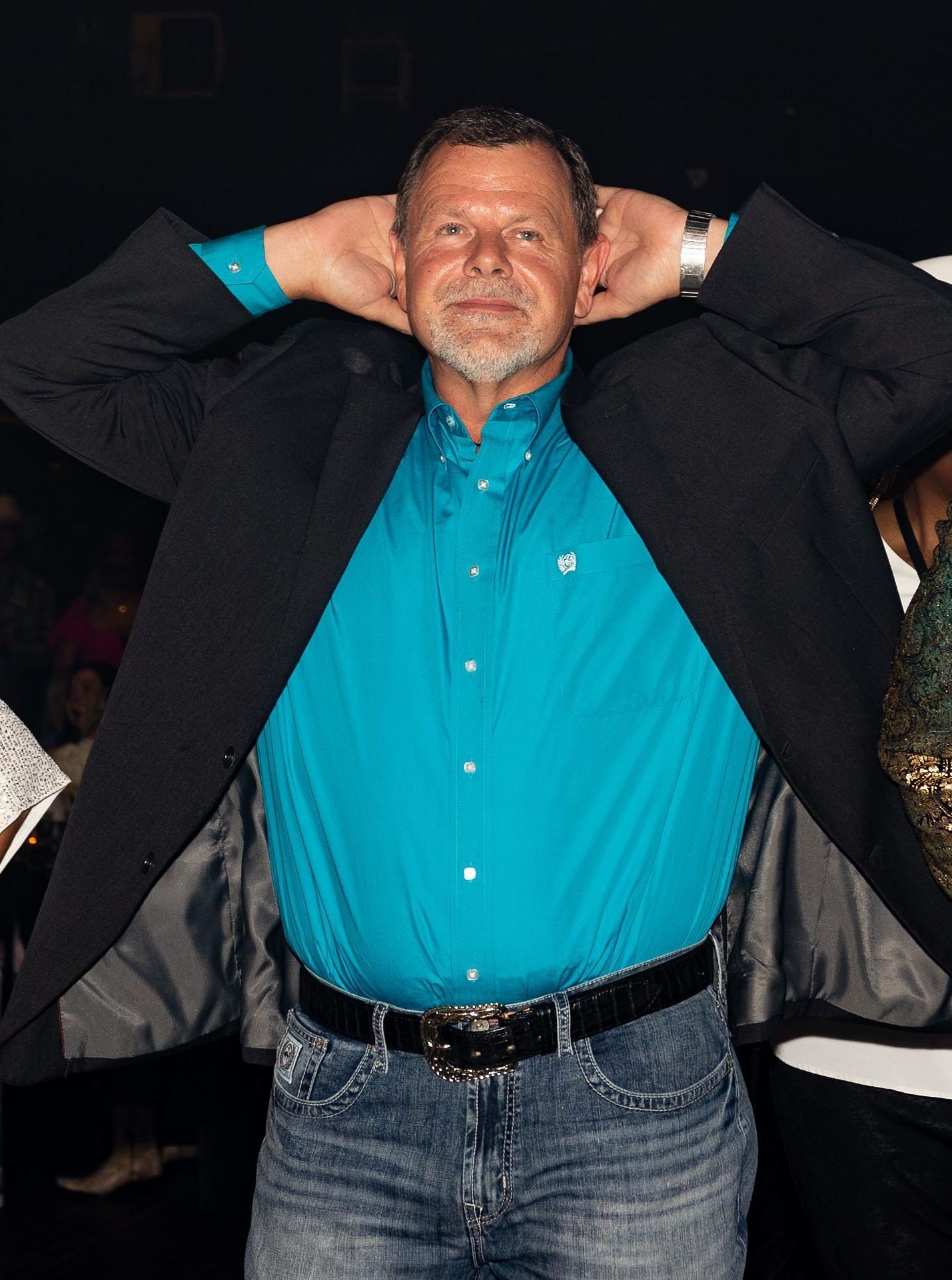 Man in blue shirt, jeans, and blazer with hands behind head, smiling.