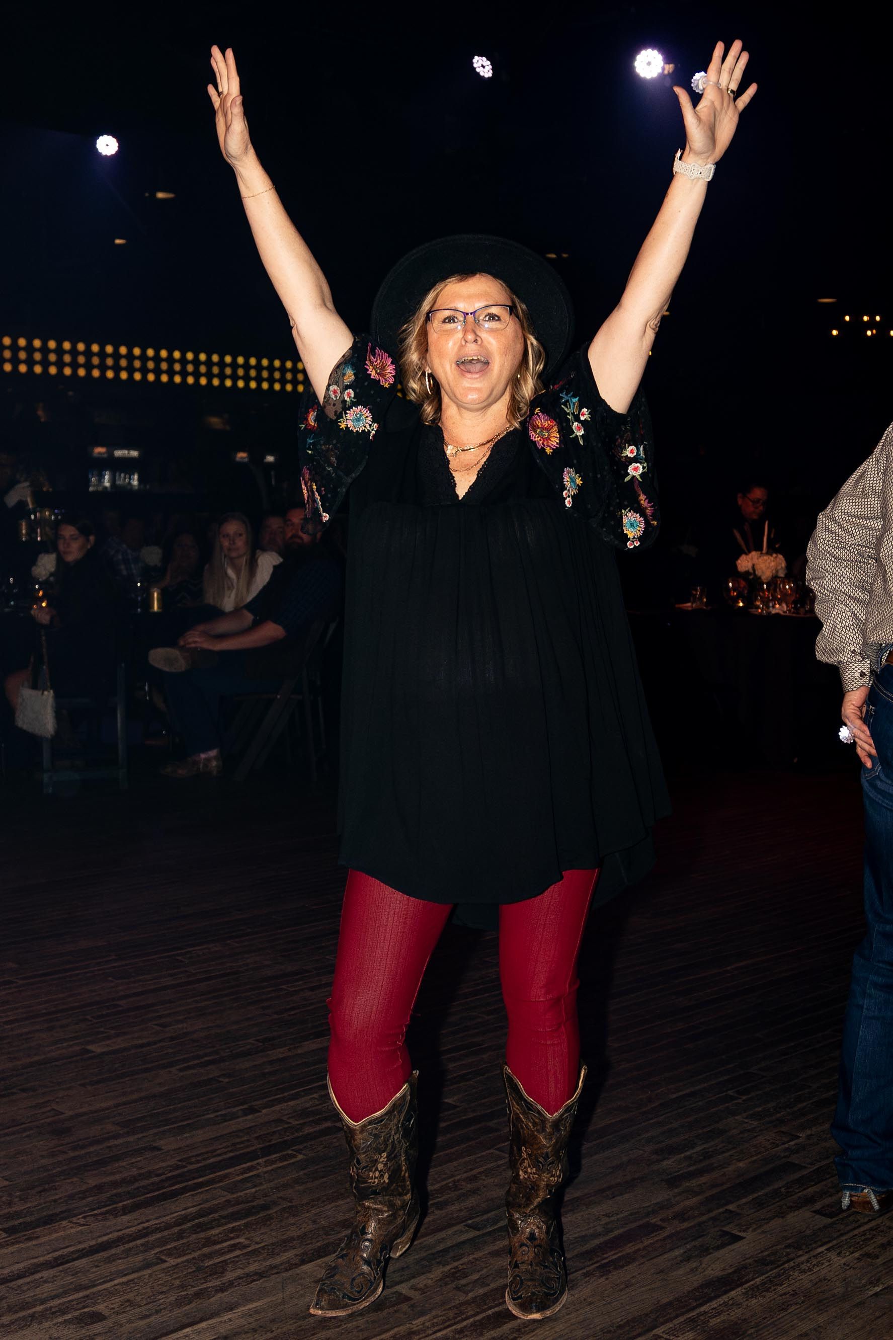 Woman in cowboy boots and red leggings raises arms in a dark venue.