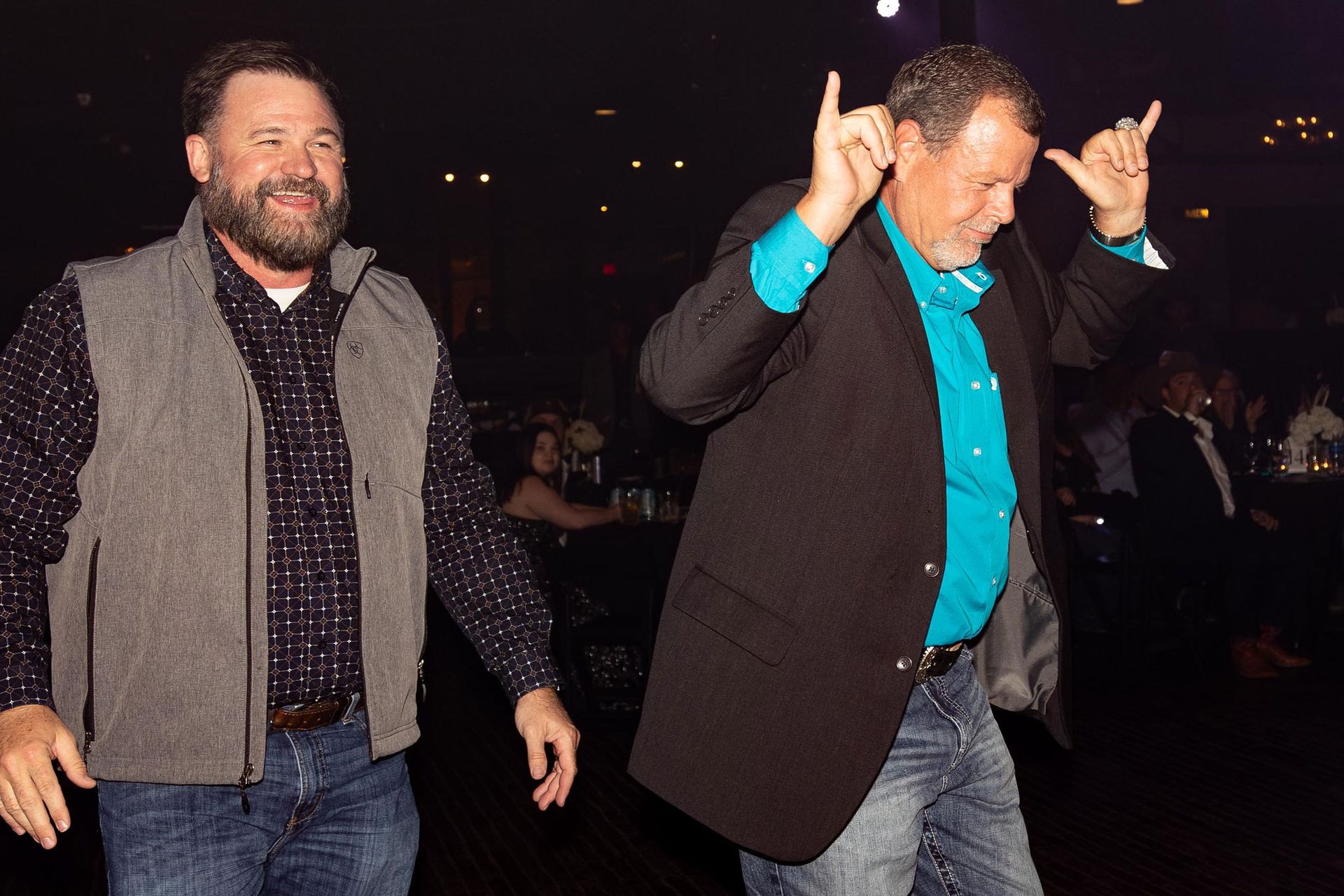 Two men dancing at an event: one in a vest, the other in a blazer, making rock-on hand gestures.