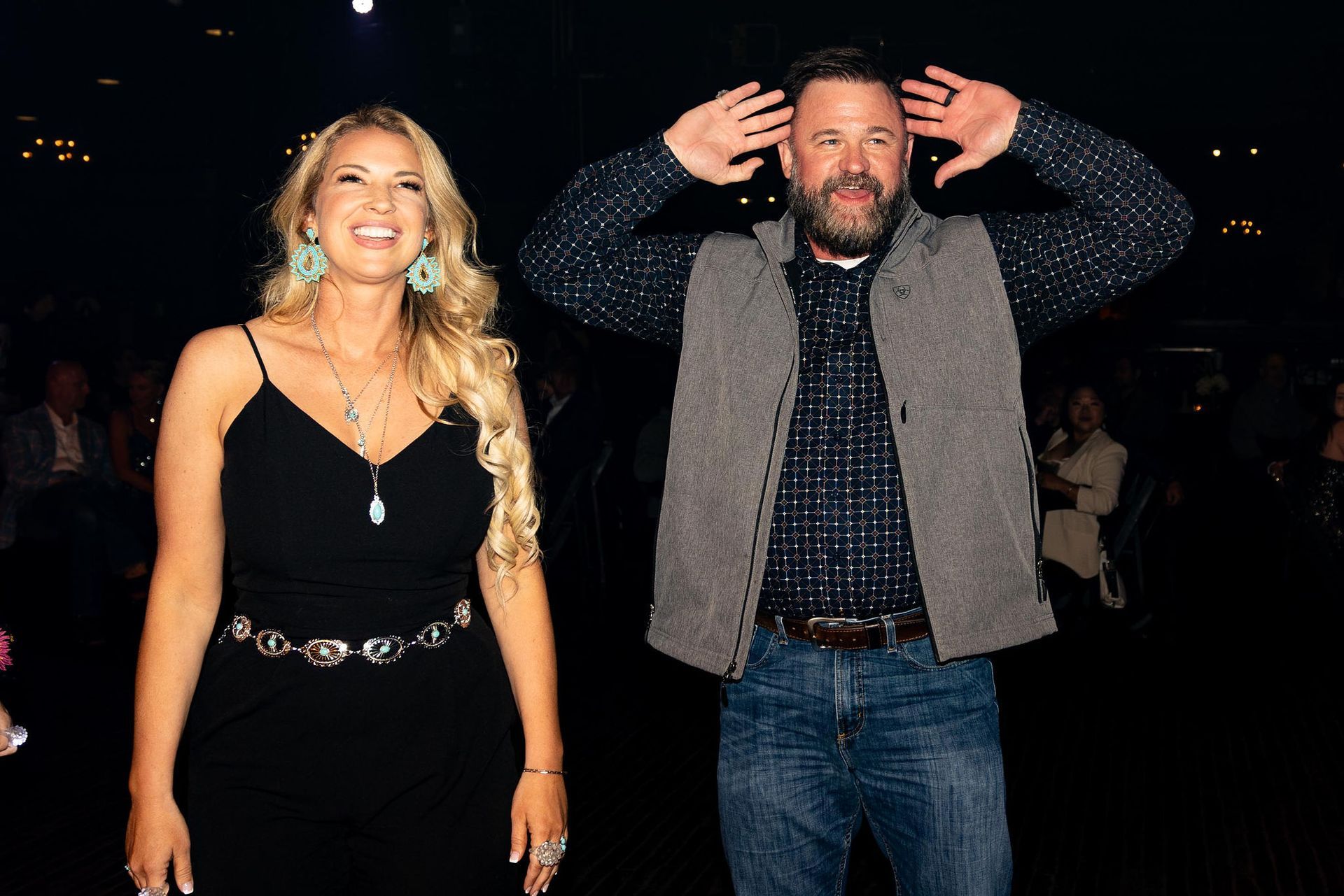Woman in black dress and man in vest pose smiling at event.