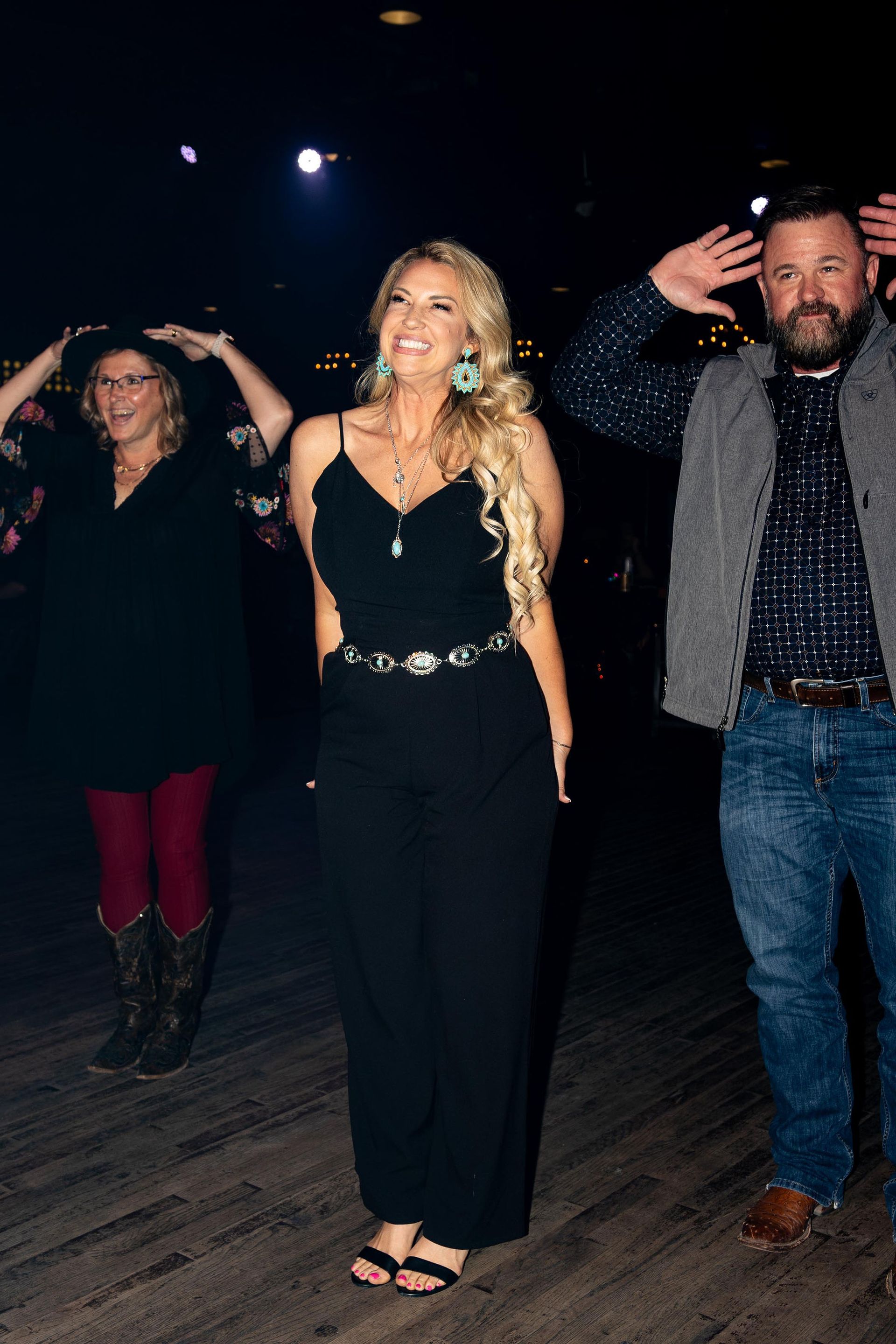 Three people dancing. Woman in black jumpsuit smiles, two others in background with hands raised. Dimly lit setting.