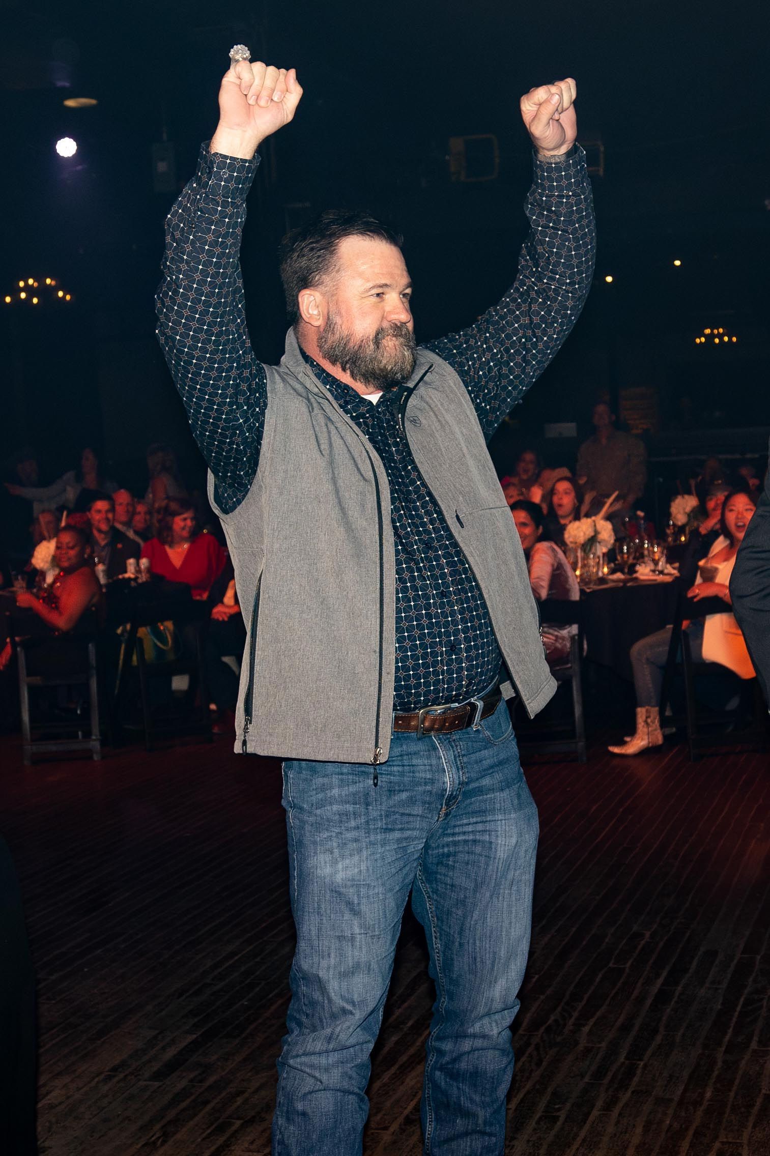 Man in jeans and vest raises arms in celebration at a large event.