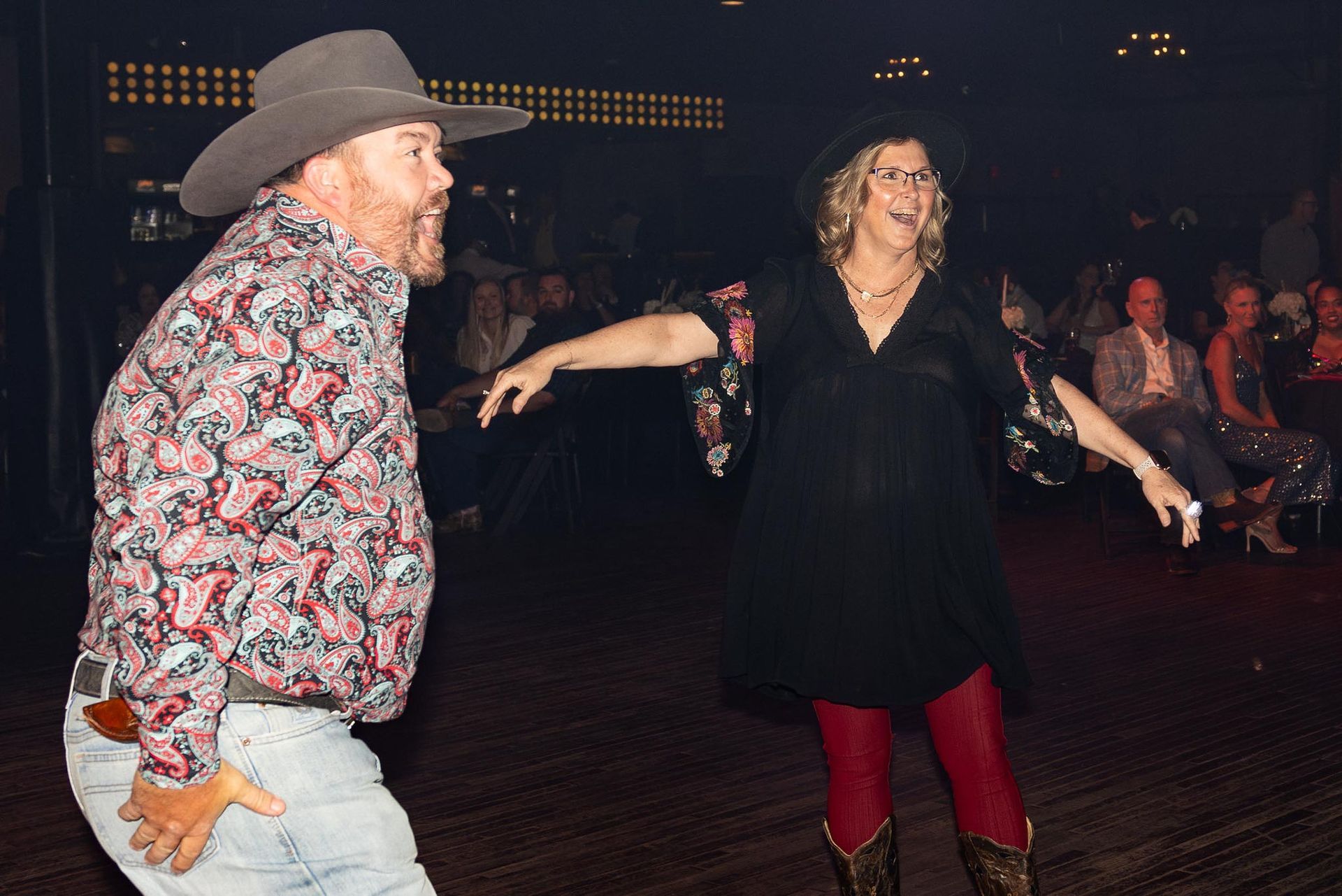 Man in cowboy hat and woman in hat dance at event, both smiling.