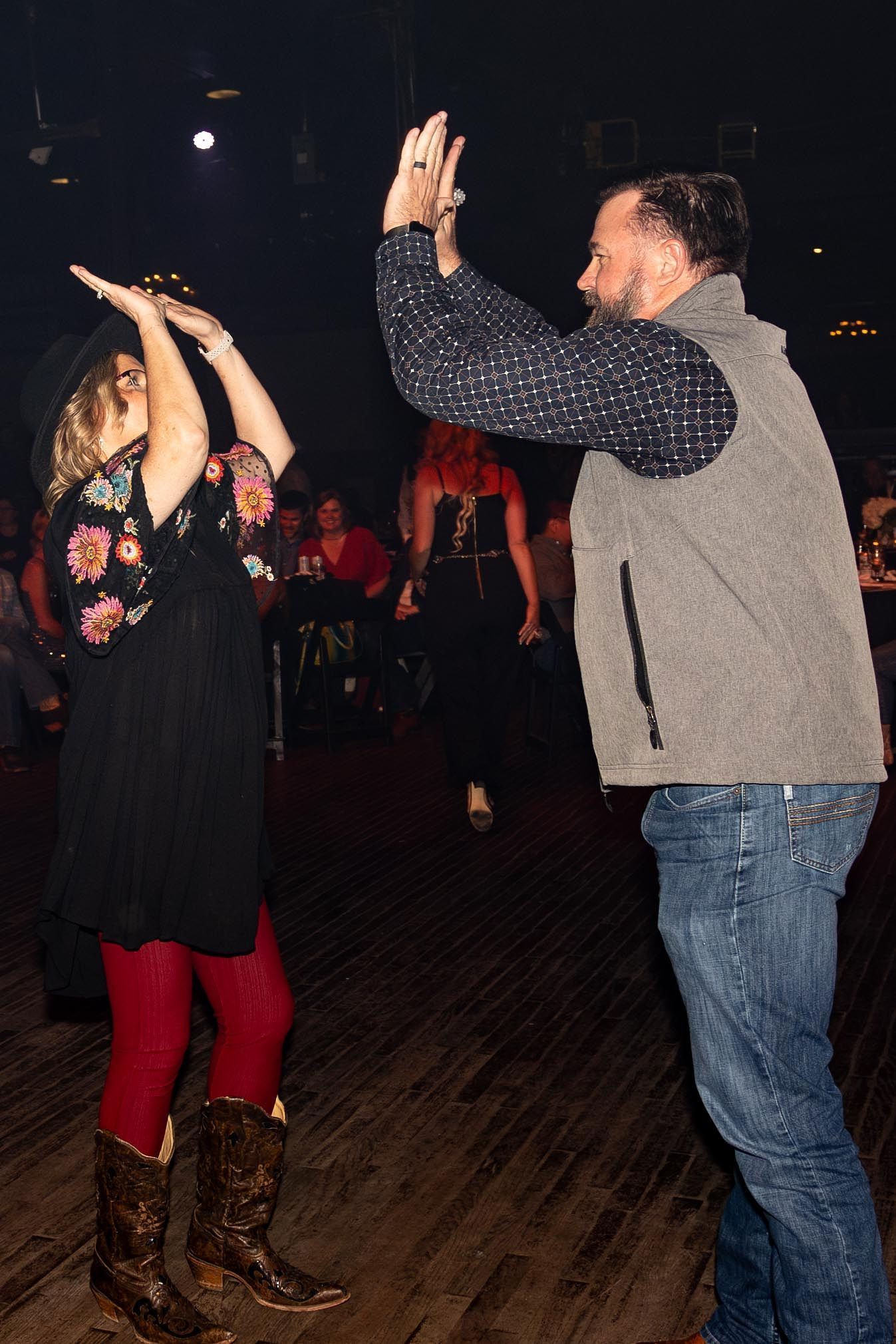Woman in black dress and hat, red tights, boots, high-fiving a man in jeans and vest on a dance floor.