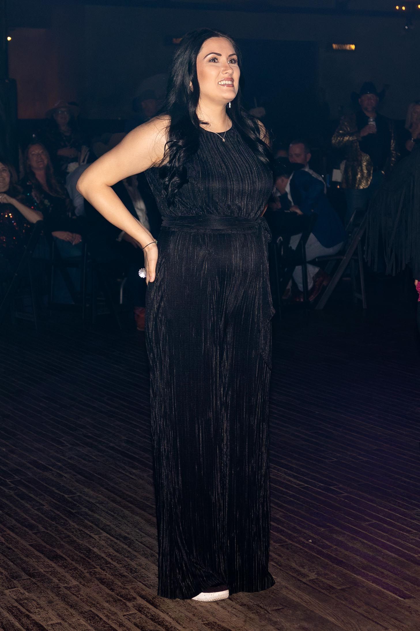 Woman in a black gown at an event, arm on hip, looking to the left. Dimly lit.