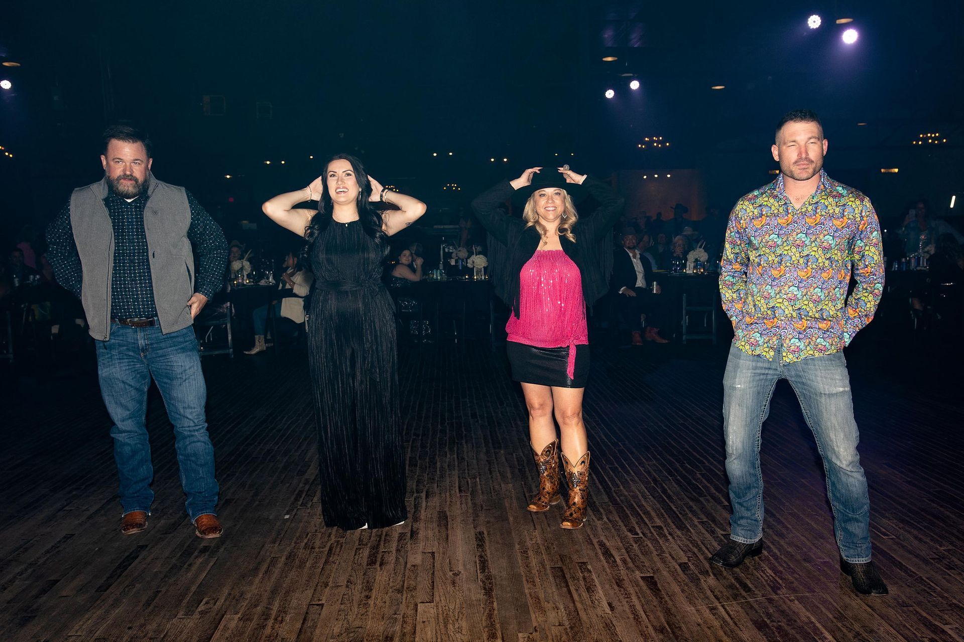 Four people posing on a stage. Two women and two men with various outfits and expressions. Dark background.