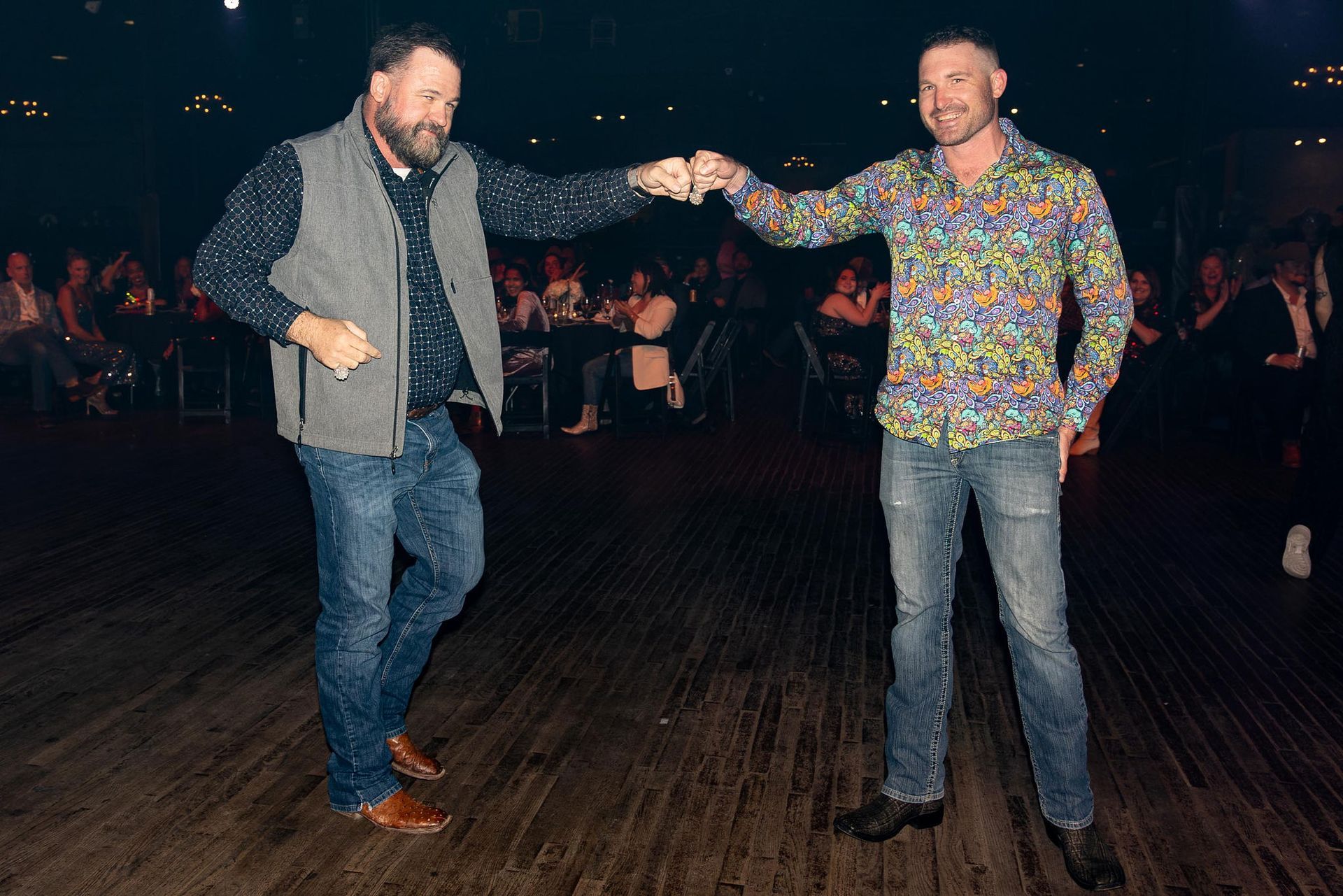 Two men dancing on a wooden floor at an event. One fist bumps, smiles. One in a vest, the other in a colorful shirt.