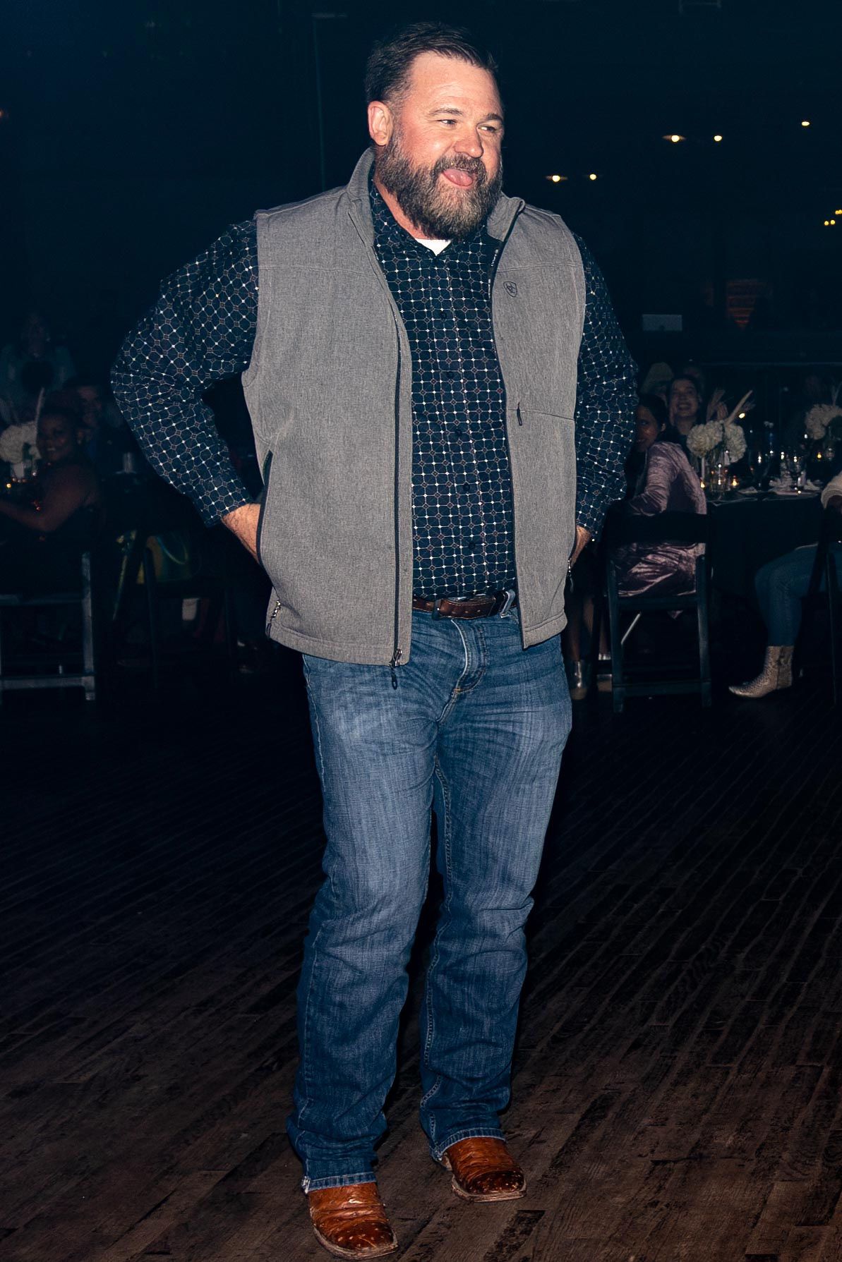 Man in a vest, jeans, and boots, hands in pockets, smiling, standing on a wooden floor, indoor event.
