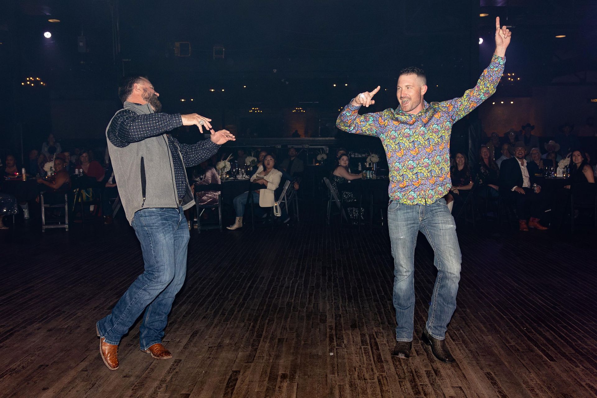 Two men dancing on a wooden floor in front of seated audience. One raises a hand, smiling. The other imitates.