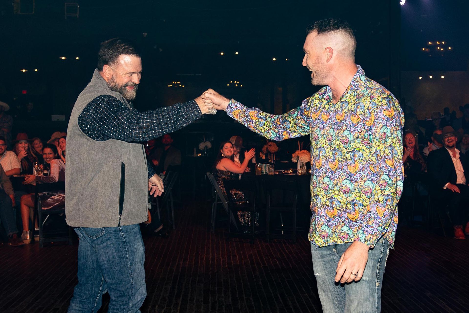 Two men fist bump at an event. One in floral shirt, the other in a vest and sparkly shirt.