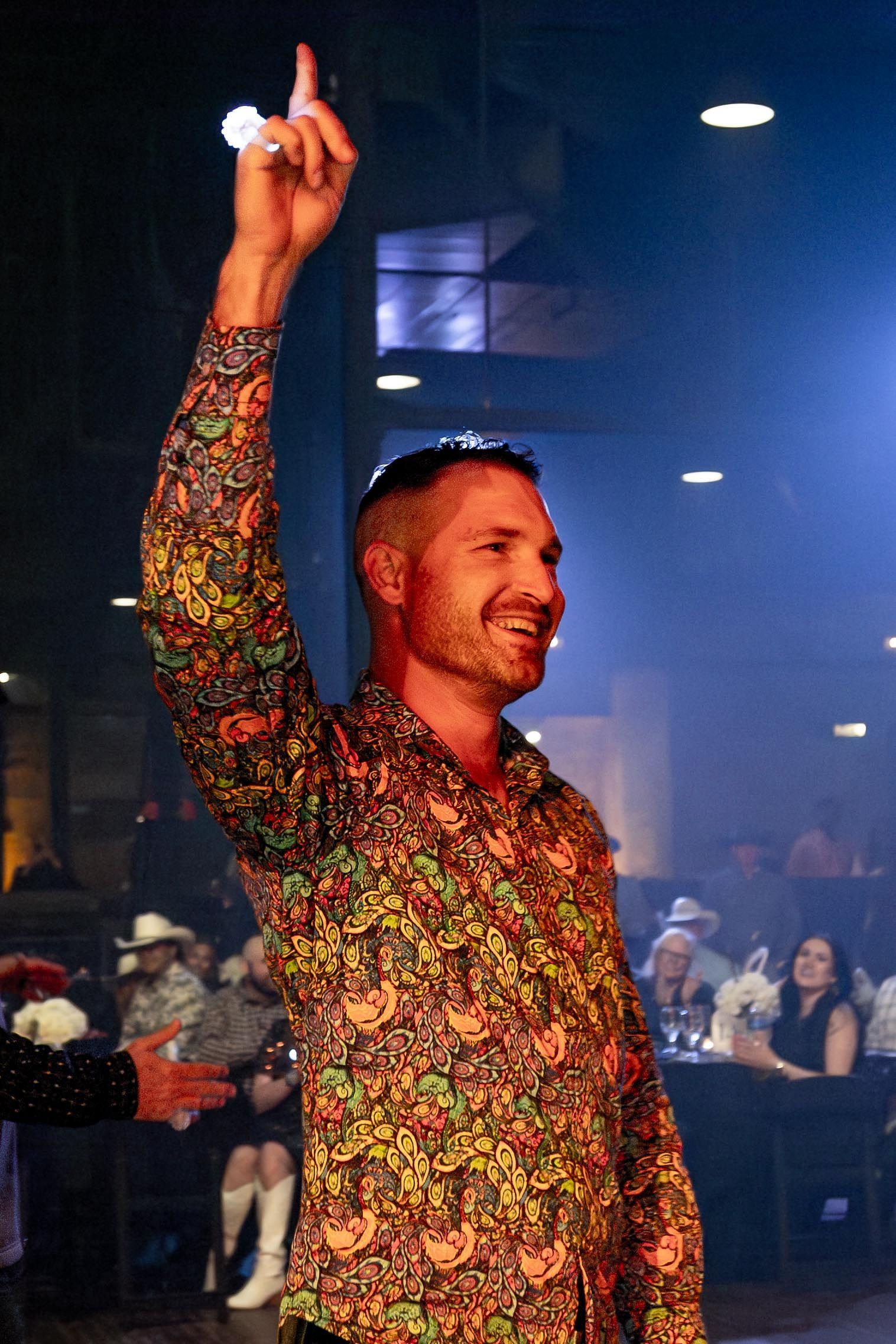 Man in colorful shirt raises arm, smiling. Inside, tables, lights.