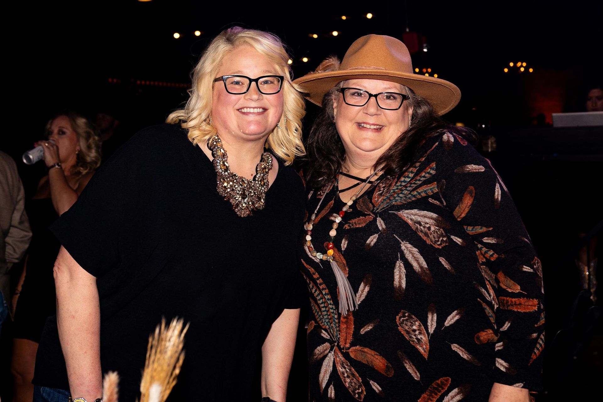 Two women smiling, one with a hat, glasses, and printed shirt, the other with a black shirt and necklace.