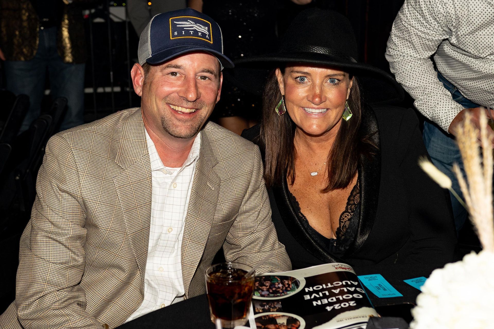 Man in a blazer and a woman in a black hat smile at the camera at a gala event.