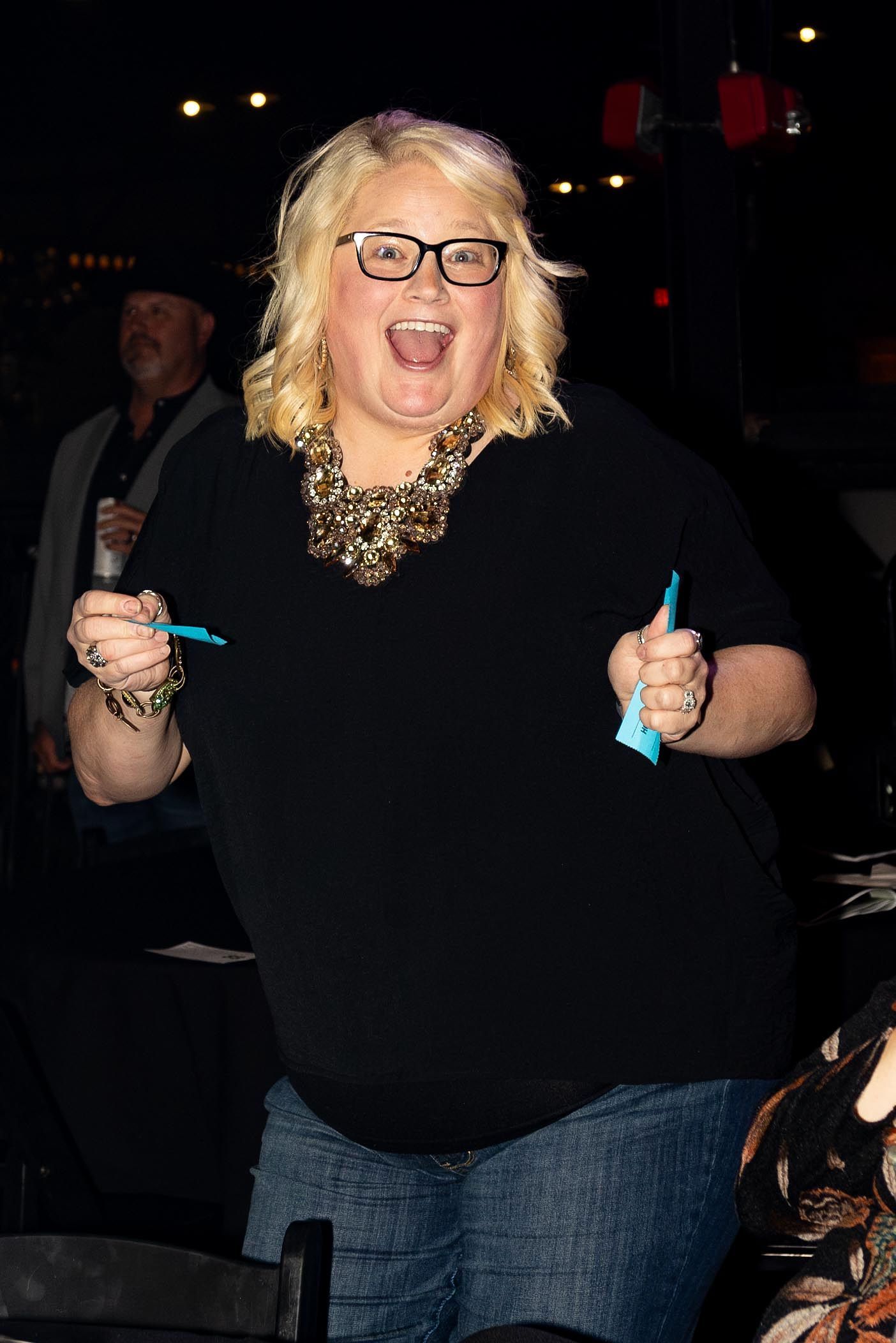 Woman with blonde hair, smiling widely, holding items, inside a dark venue.