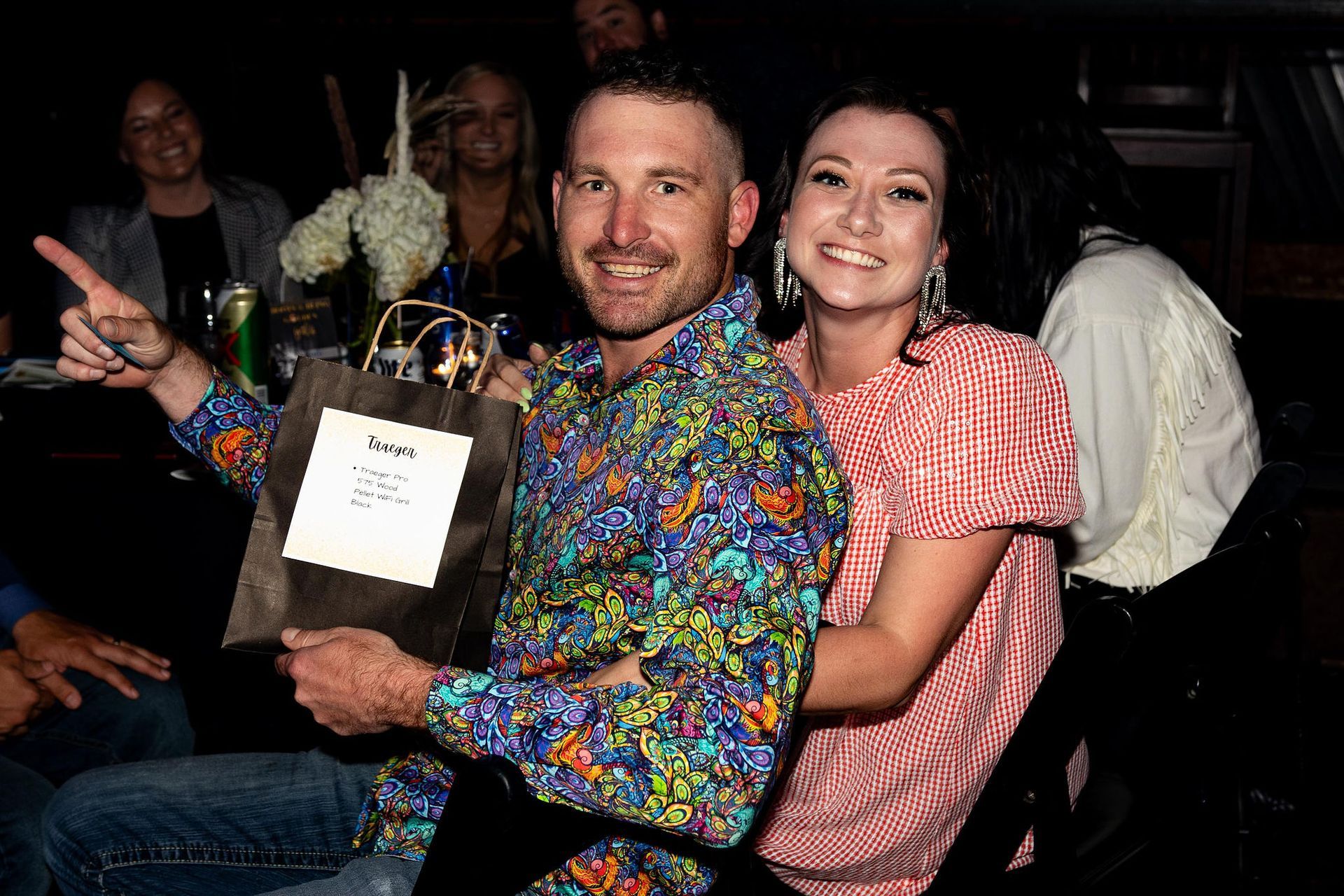 Man in colorful shirt and woman smile, holding a gift bag indoors; others in background.