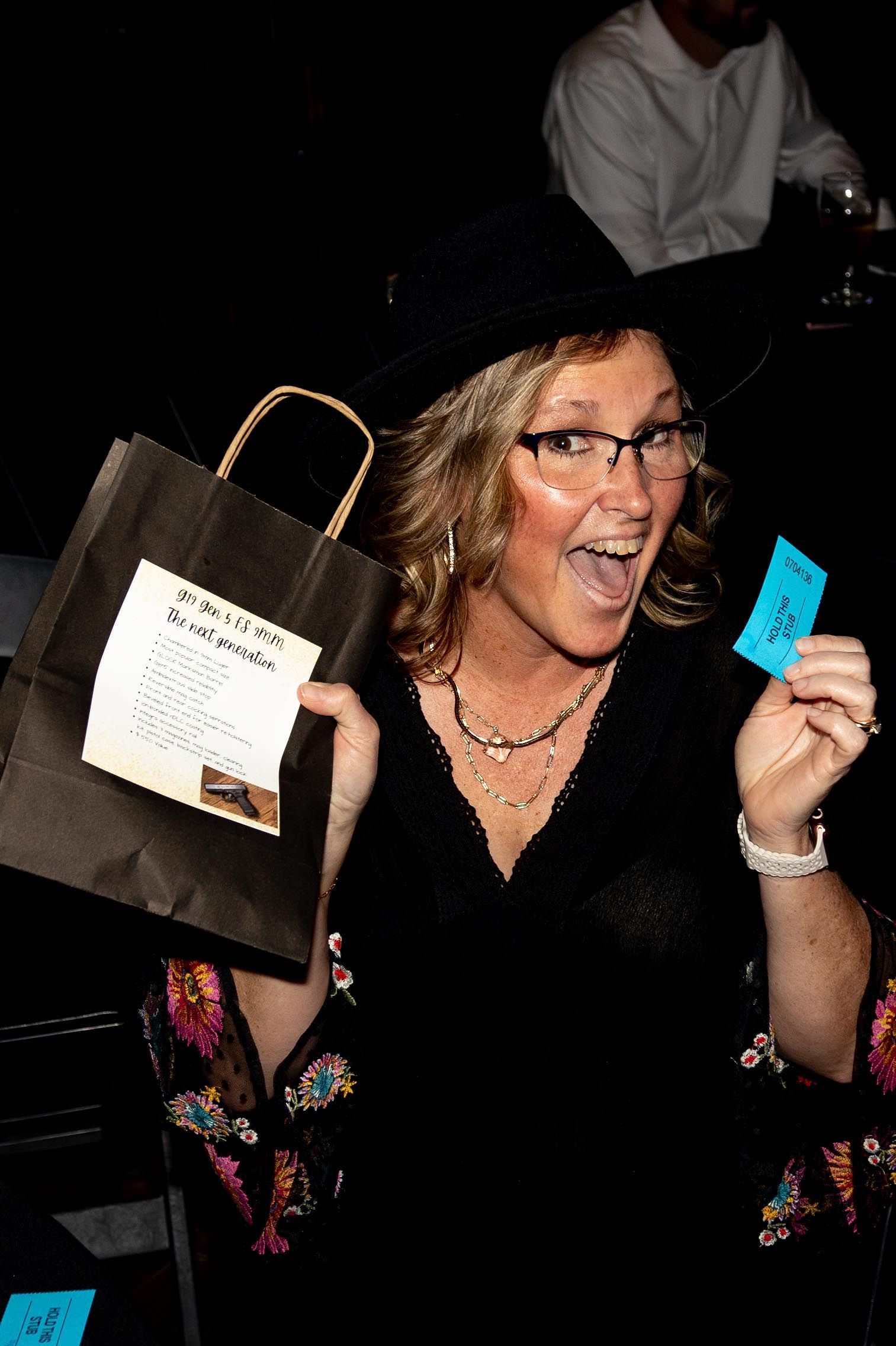 Woman in hat and glasses holds prize bag and winning ticket, excited expression.