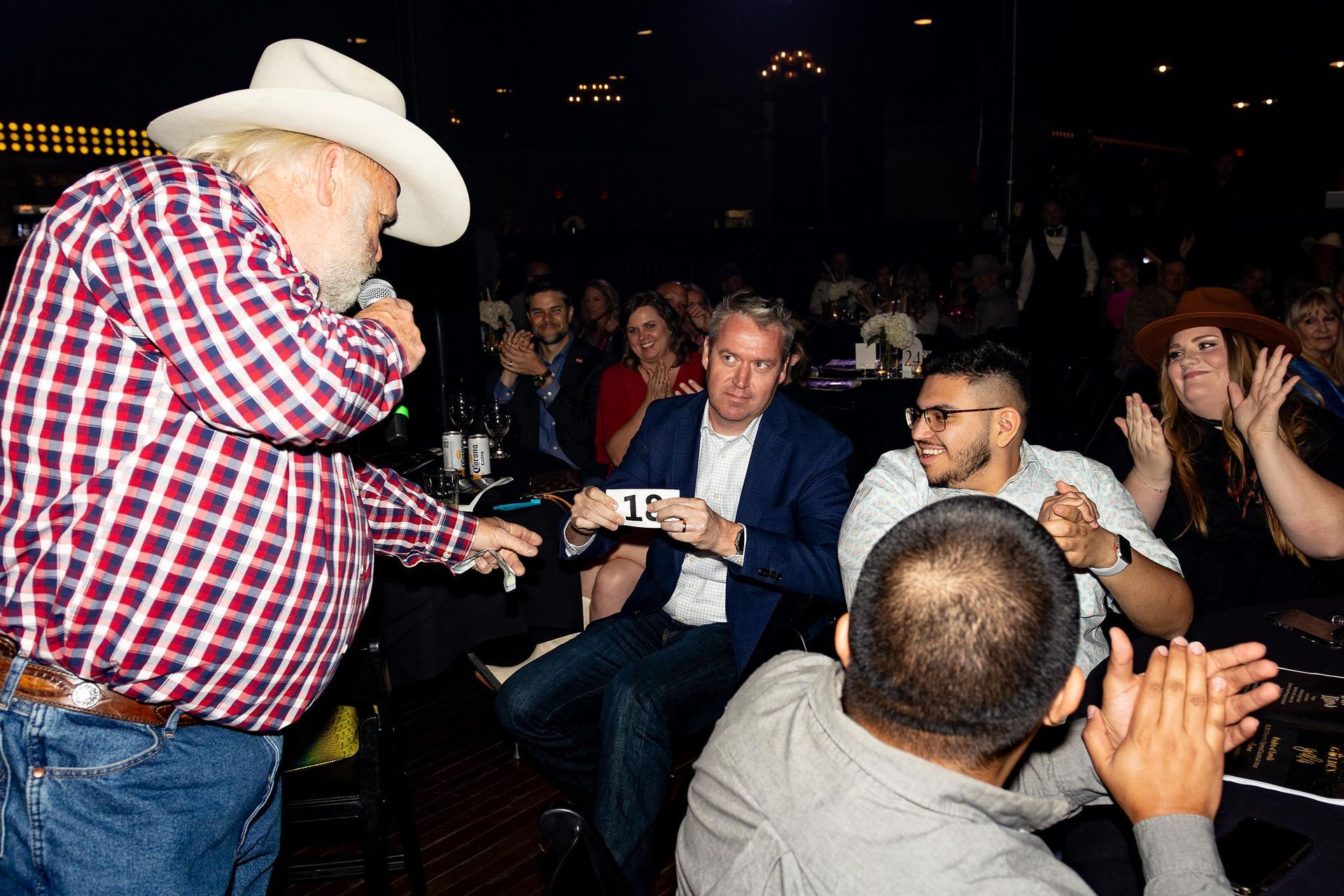 Man in cowboy hat and checkered shirt leading a lively auction; people in audience raise paddles and cheer.