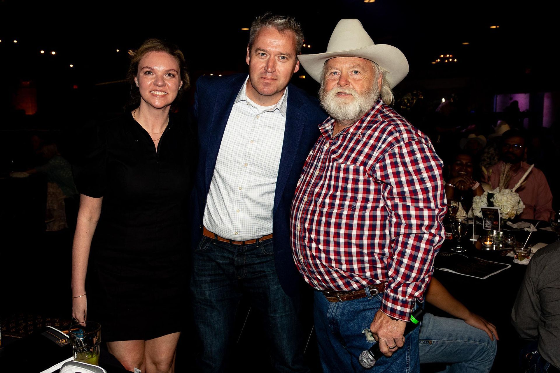 Three people pose indoors: a woman in black, a man in a blazer, and a man in a cowboy hat and plaid shirt.