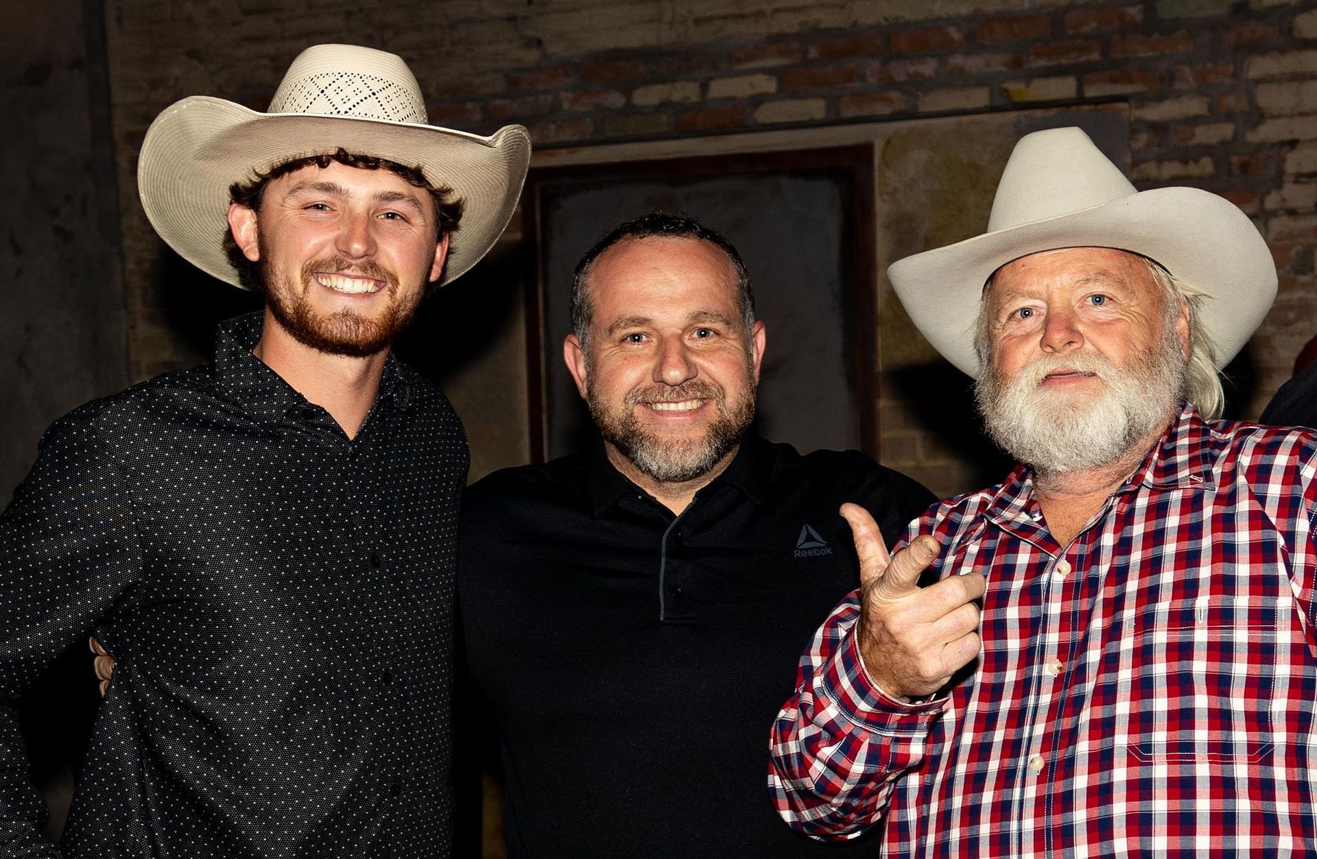 Three men in cowboy hats pose smiling. One man gives a thumbs up. Dark interior.