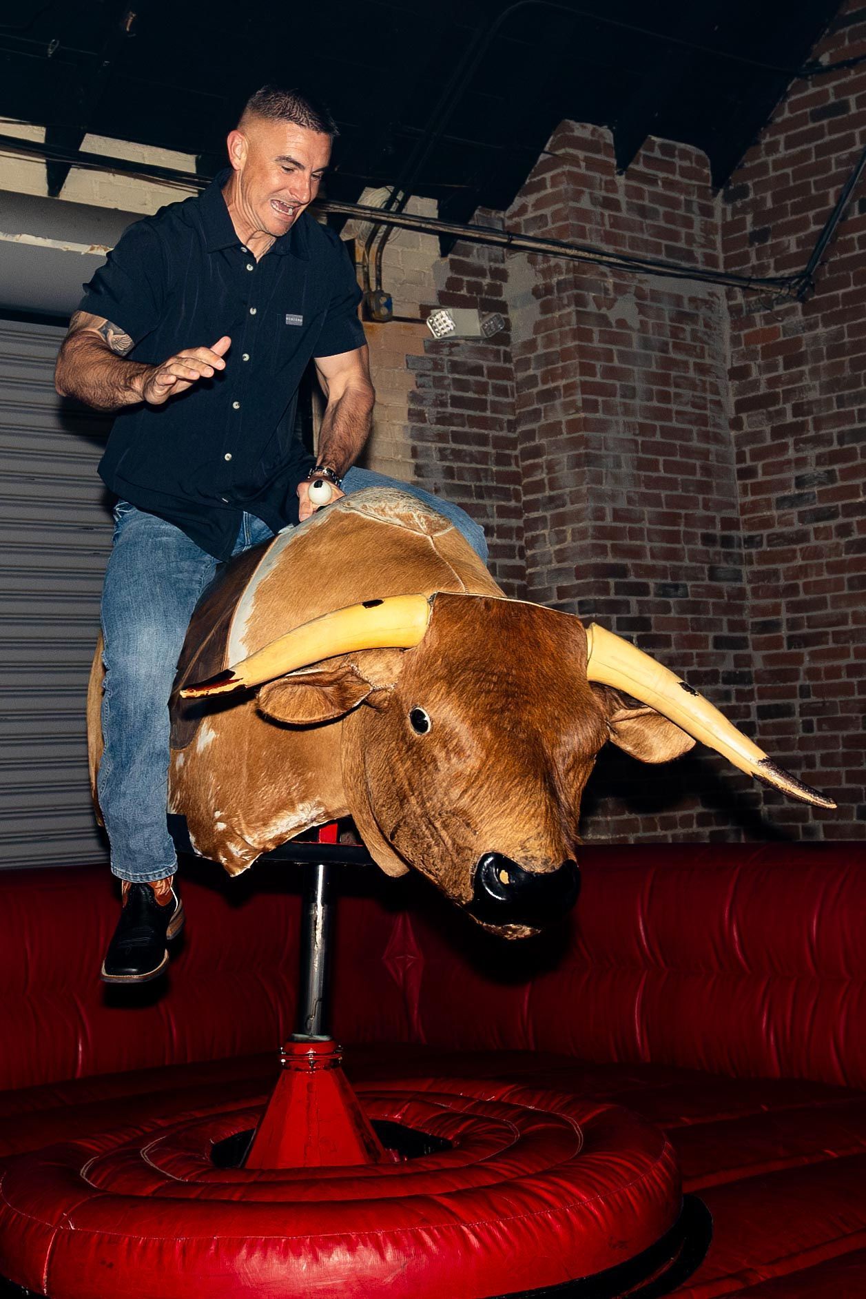 Man riding a mechanical bull at a bar; brick wall background.