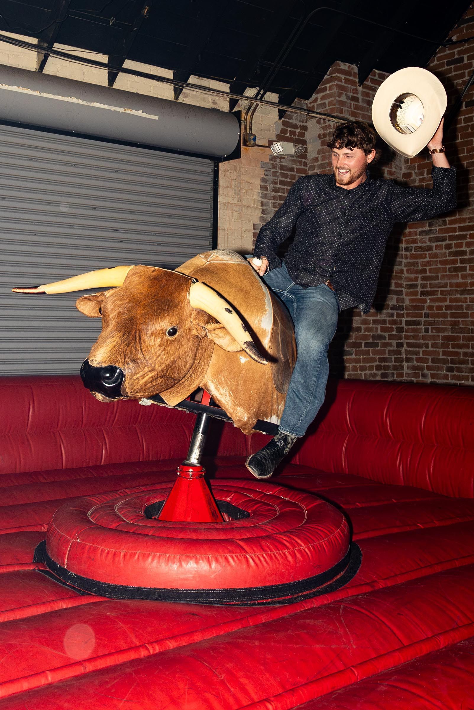 Man riding a mechanical bull indoors, wearing a cowboy hat, raising his arm in excitement. Red padded area.