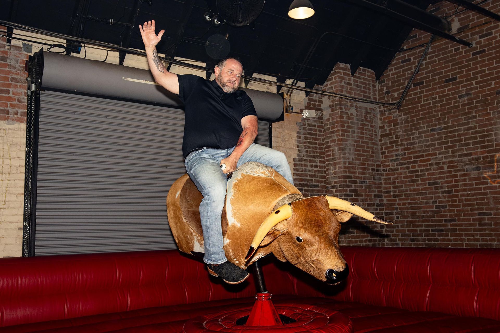 Man riding mechanical bull, arm raised. Red padded area, brick wall background.