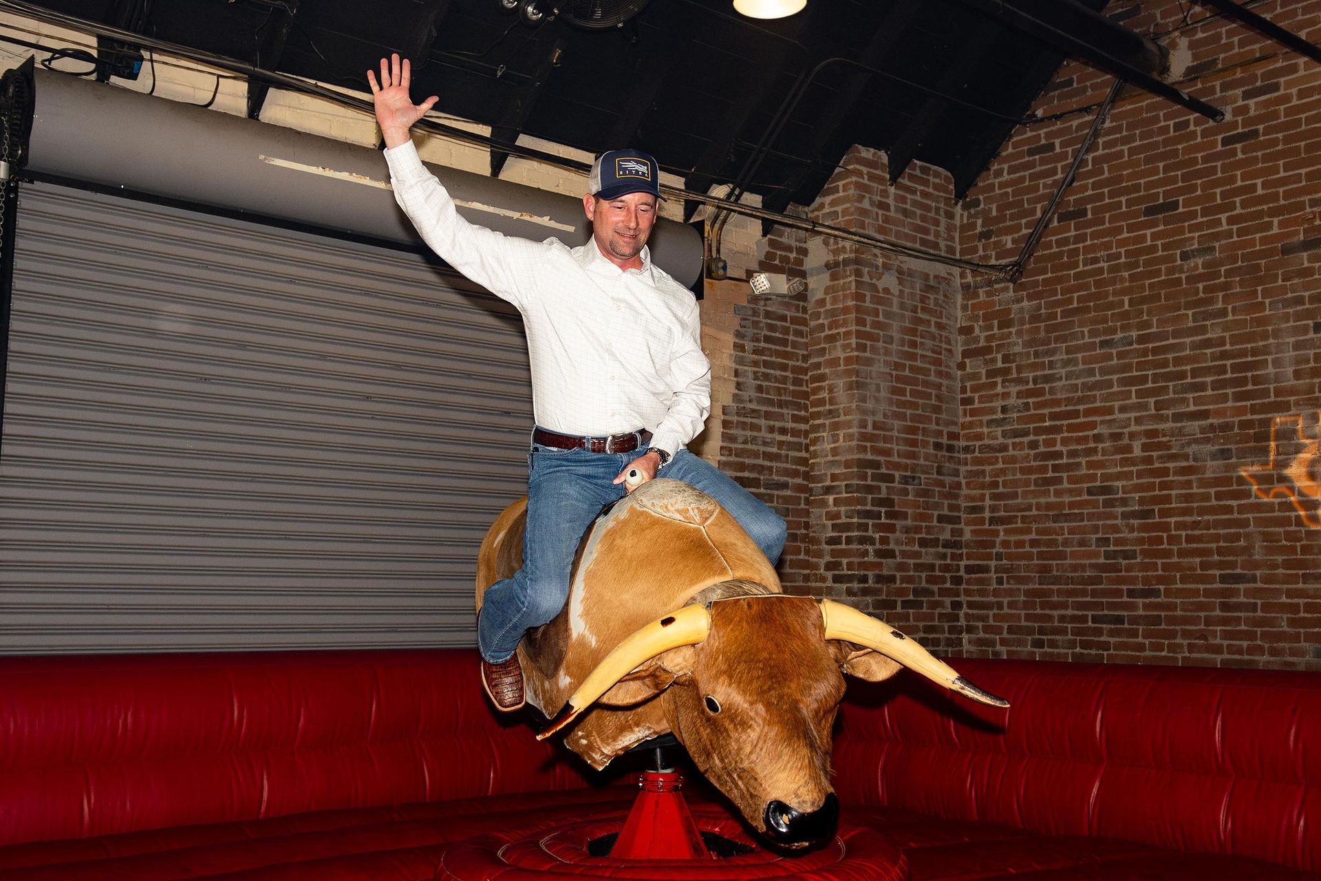 Man riding a mechanical bull, raising his hand in a bar. Red padding, brick wall.