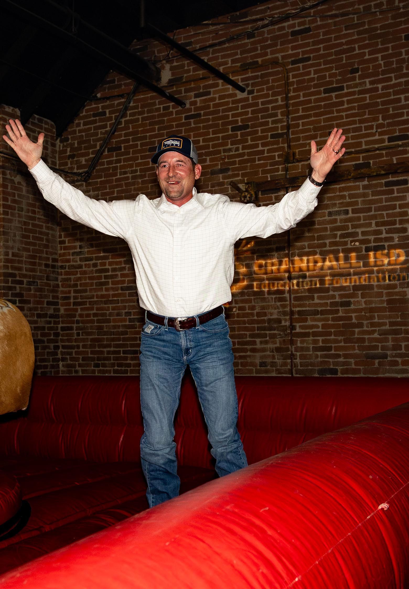 Man with arms outstretched, smiling, in blue jeans and white shirt, on red inflatable. Brick wall background.
