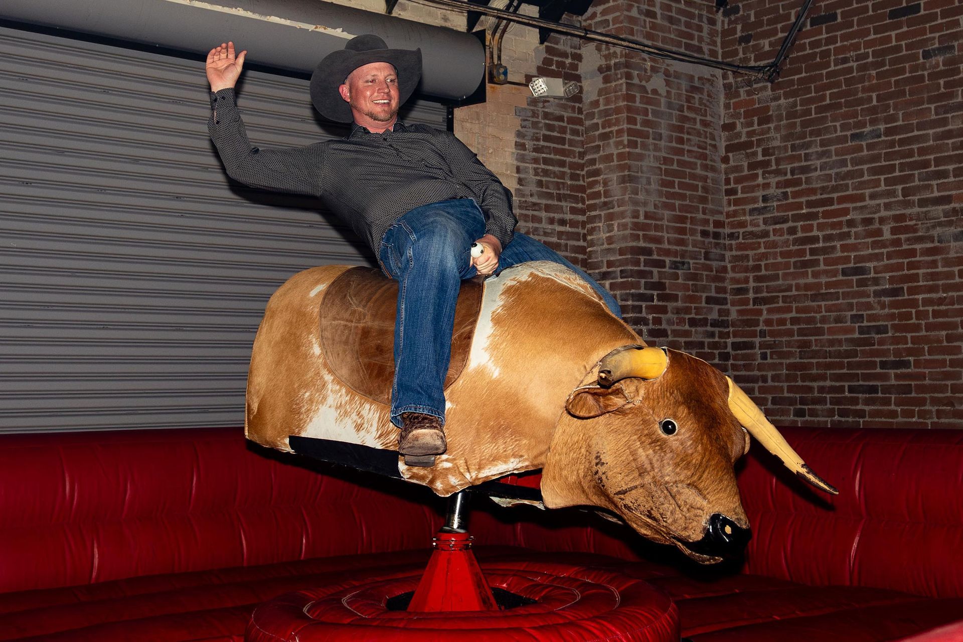 Man in cowboy hat riding mechanical bull, raising his arm in a brick-walled setting.