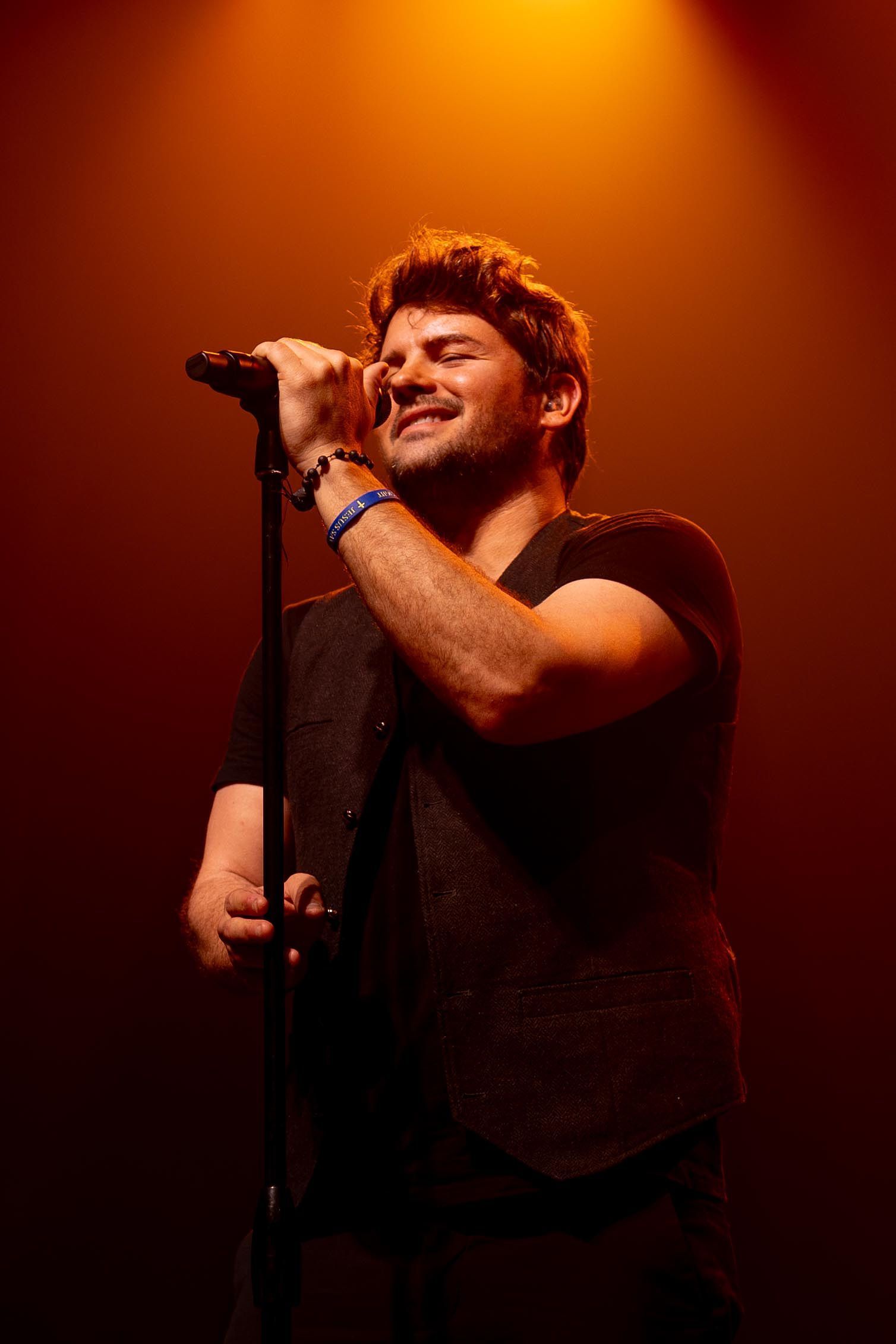 Man singing into a microphone on stage, smiling. Wearing a black vest and shirt, lit by orange stage lights.