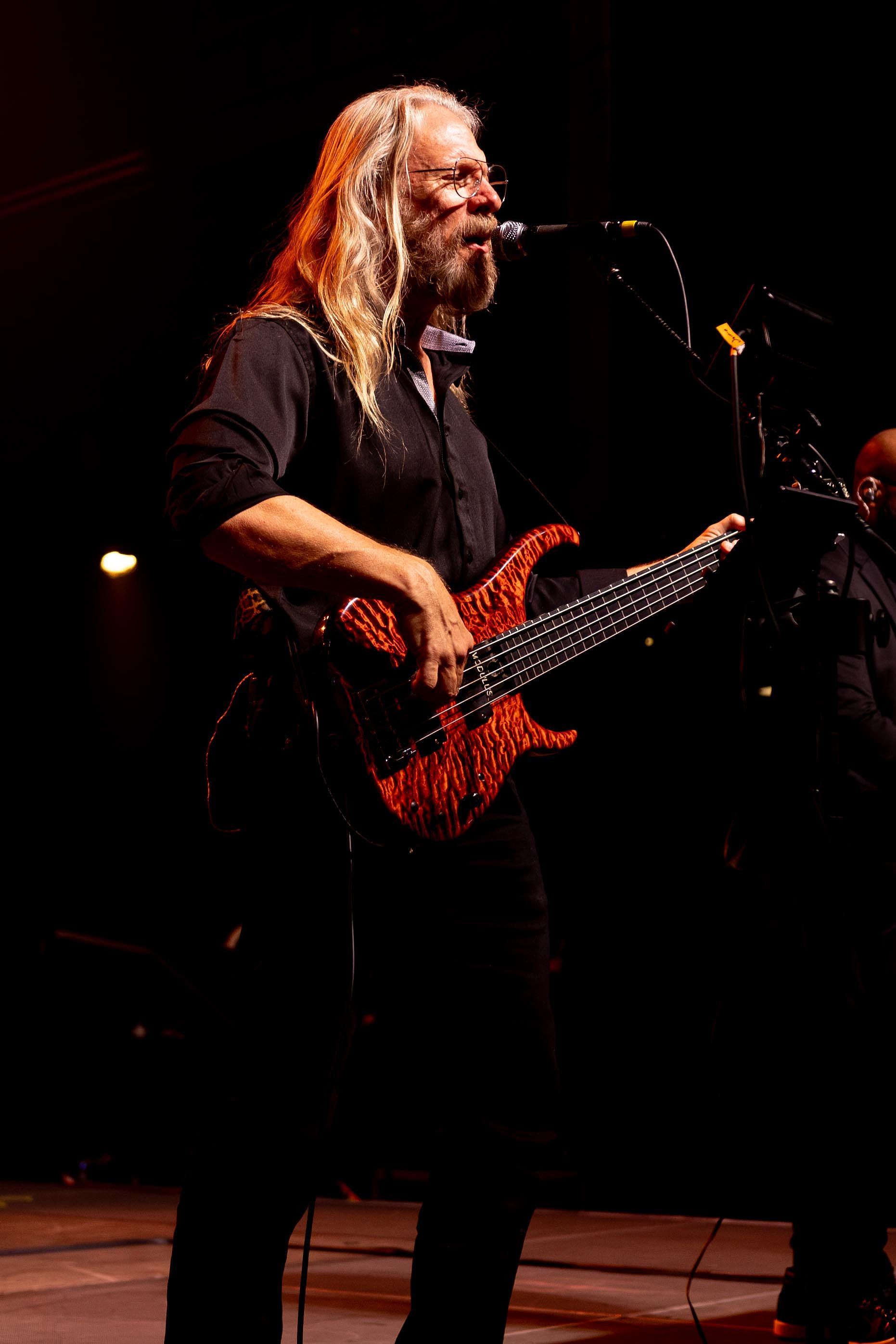 Man with long white hair plays a red bass guitar on stage, singing into a microphone. Dark setting.