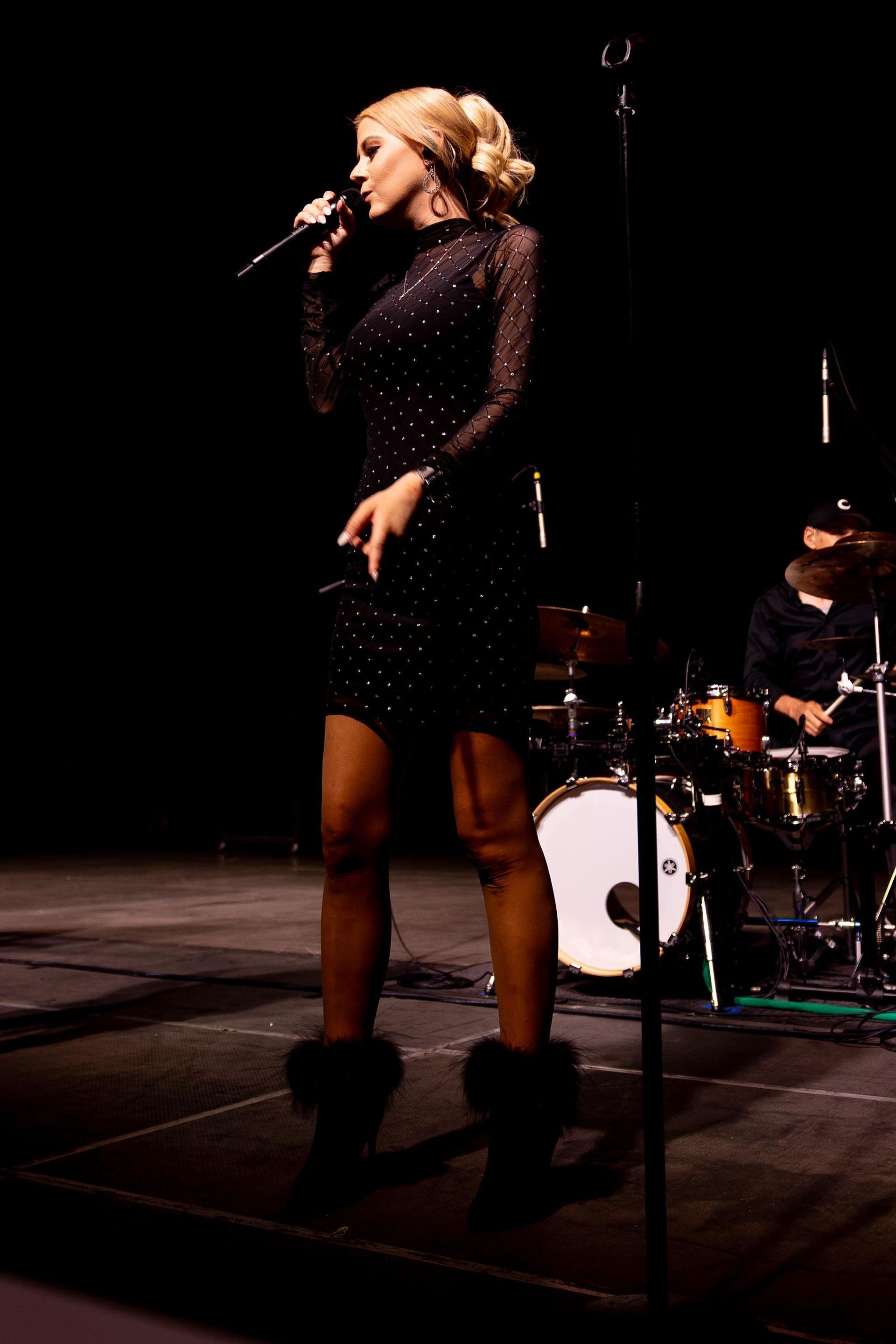 Woman in a black dress singing on stage, blonde hair up, holding a microphone. Drummer in background.