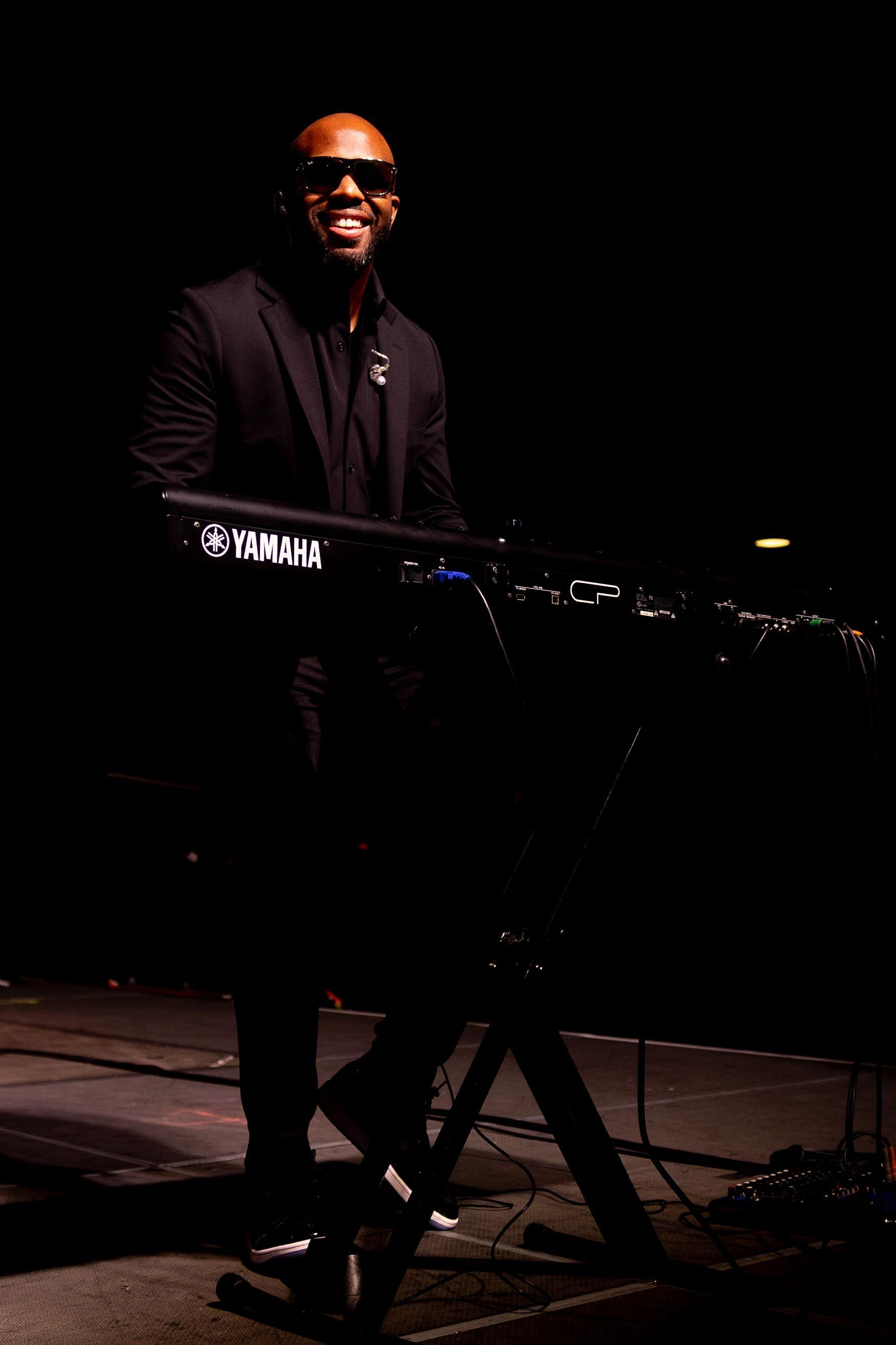 Smiling Black man playing a Yamaha keyboard on a dark stage, wearing a black suit and sunglasses.