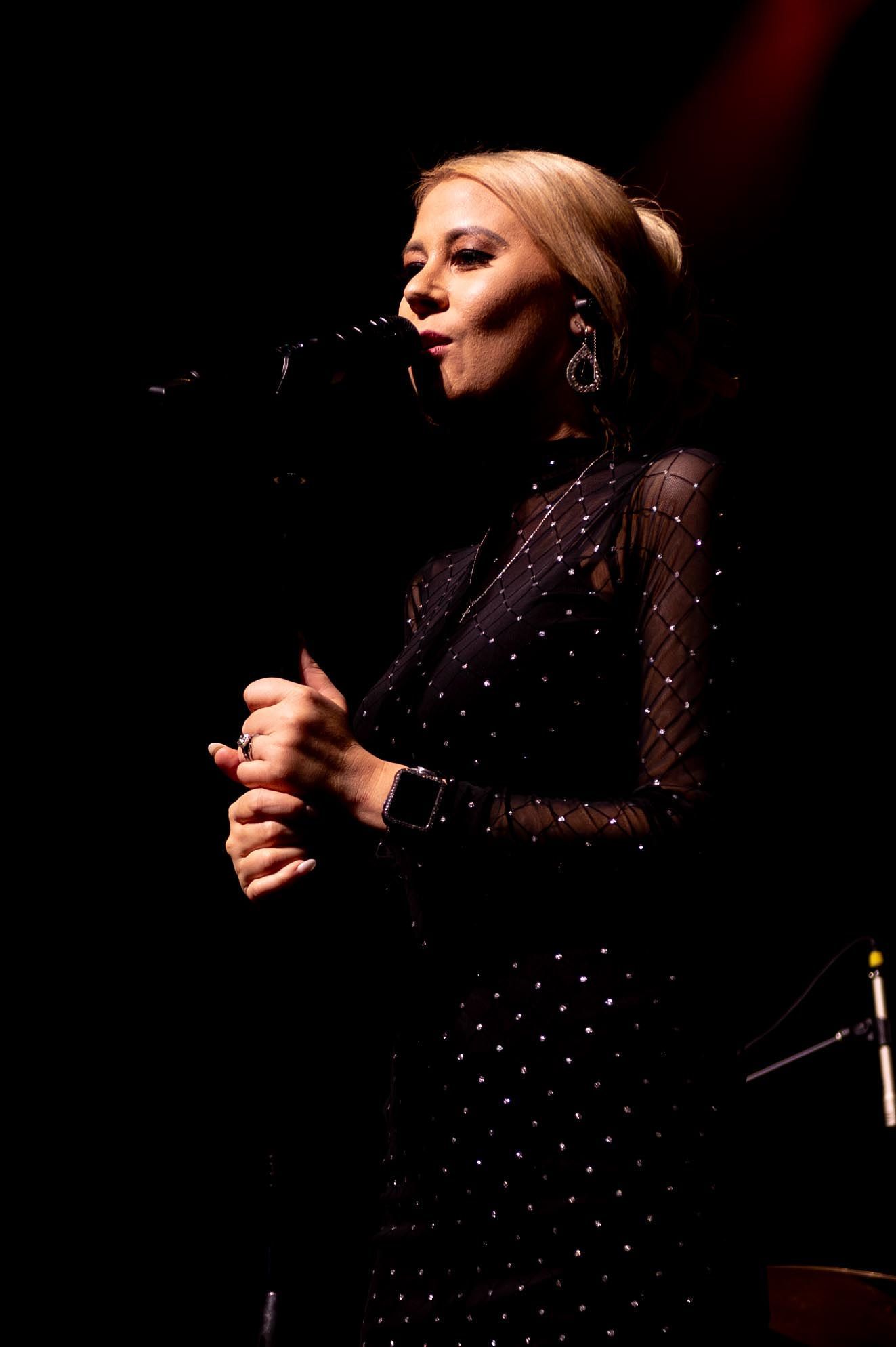 Woman with blonde hair sings into a microphone, wearing a dark dress with silver dots, on stage.