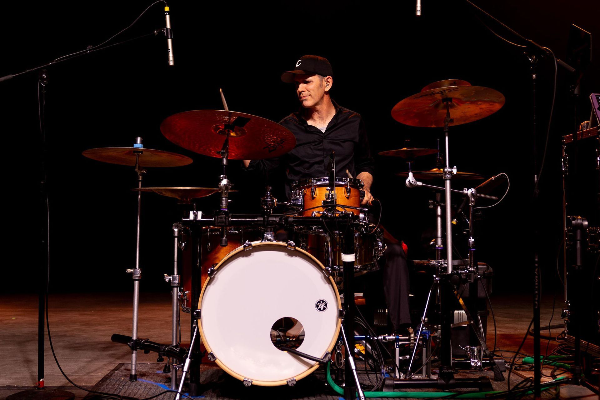 Drummer playing a drum kit on stage. He's wearing a black shirt and hat, smiling. The drum kit is surrounded by microphones.