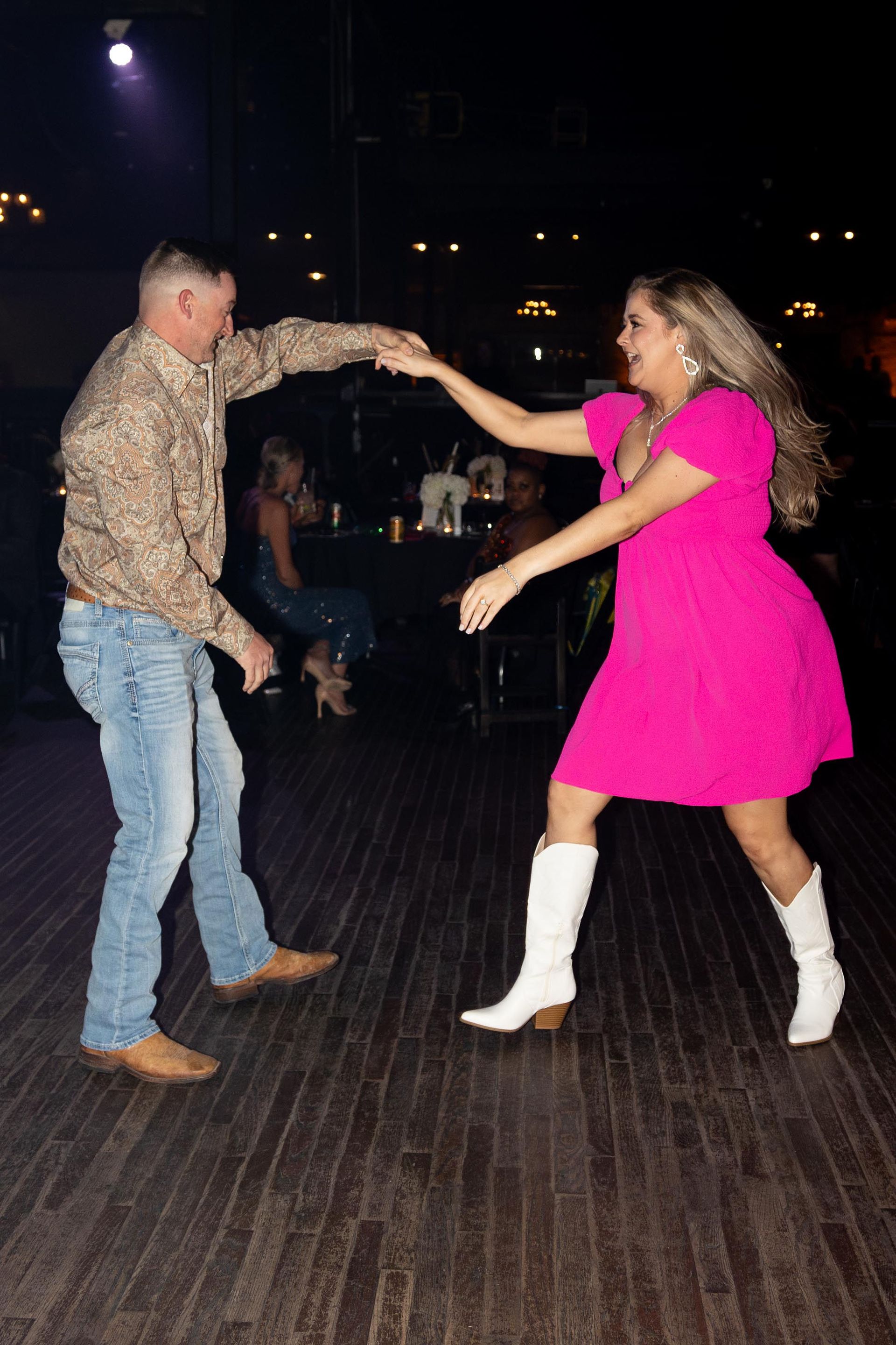 Man and woman dance. Woman in pink dress and white boots. Man in cowboy shirt and jeans. Dark room.