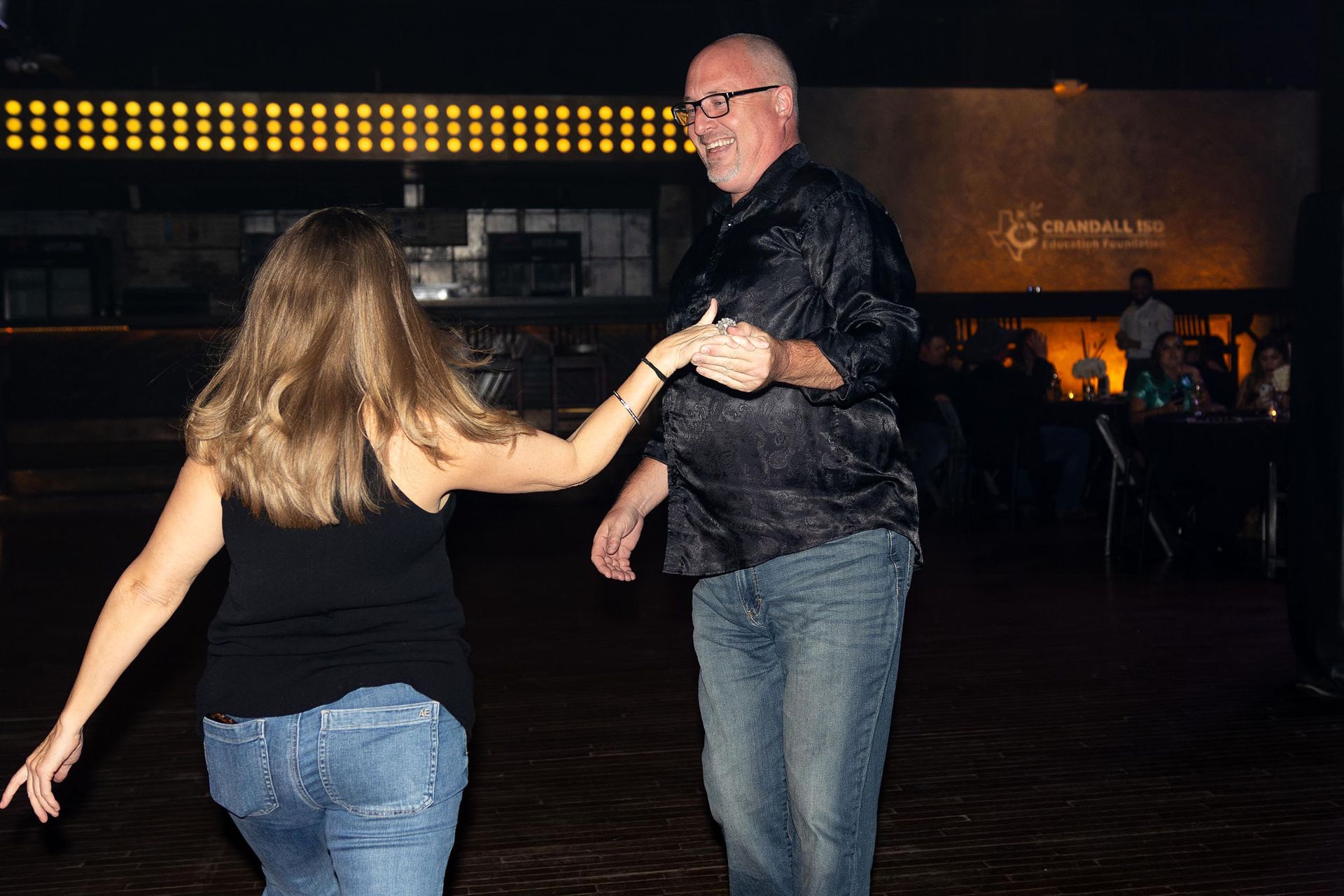 Couple dancing, man in black shirt and jeans, woman in black tank top and jeans, dim lit venue.