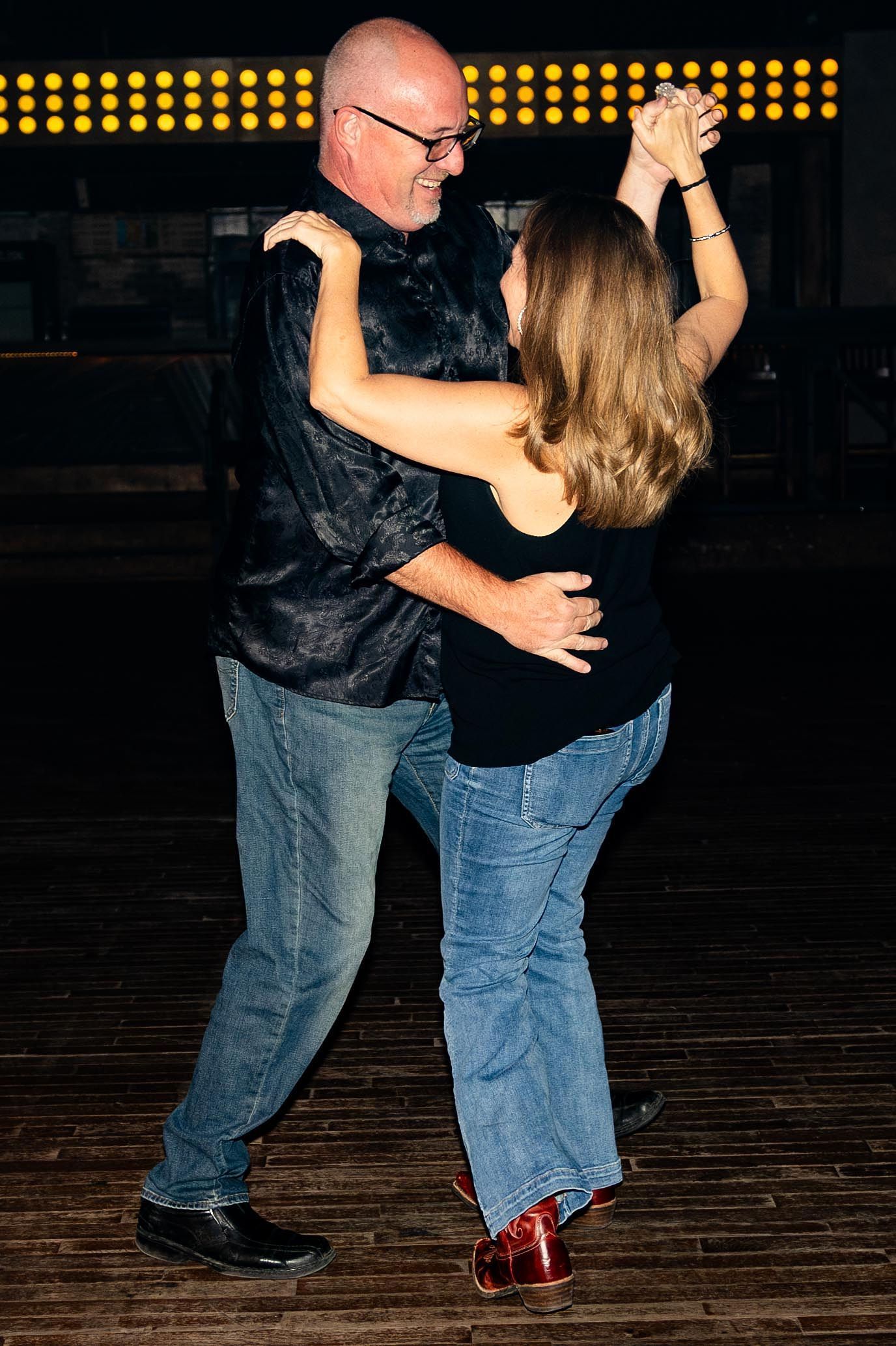 A man and woman dance in a dimly lit room. The man wears a black shirt and jeans. The woman wears a black tank top and jeans.