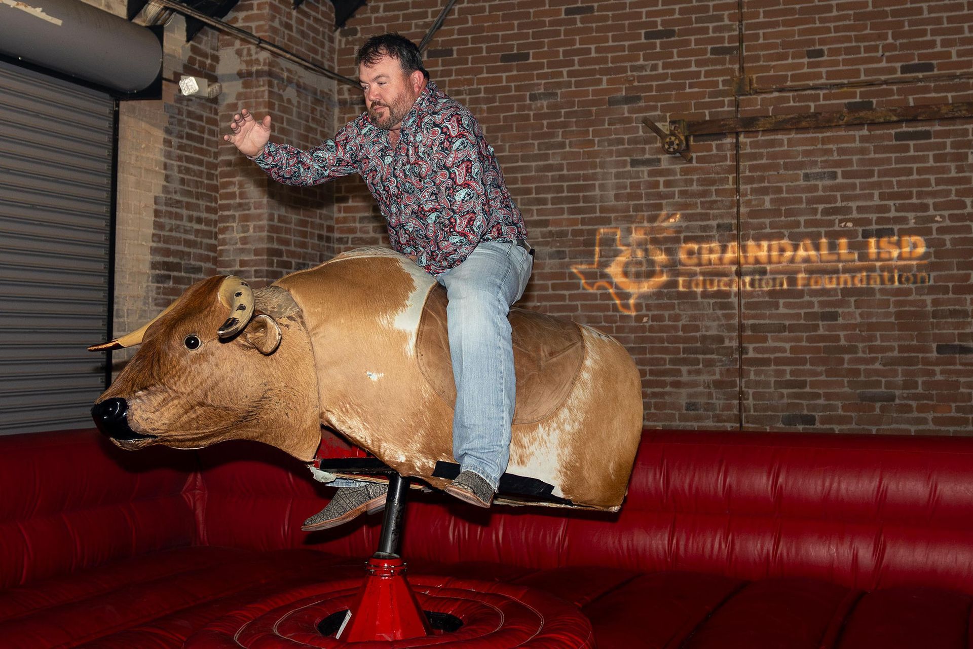 Man riding a mechanical bull indoors. Wearing patterned shirt and jeans, raising his arm. 