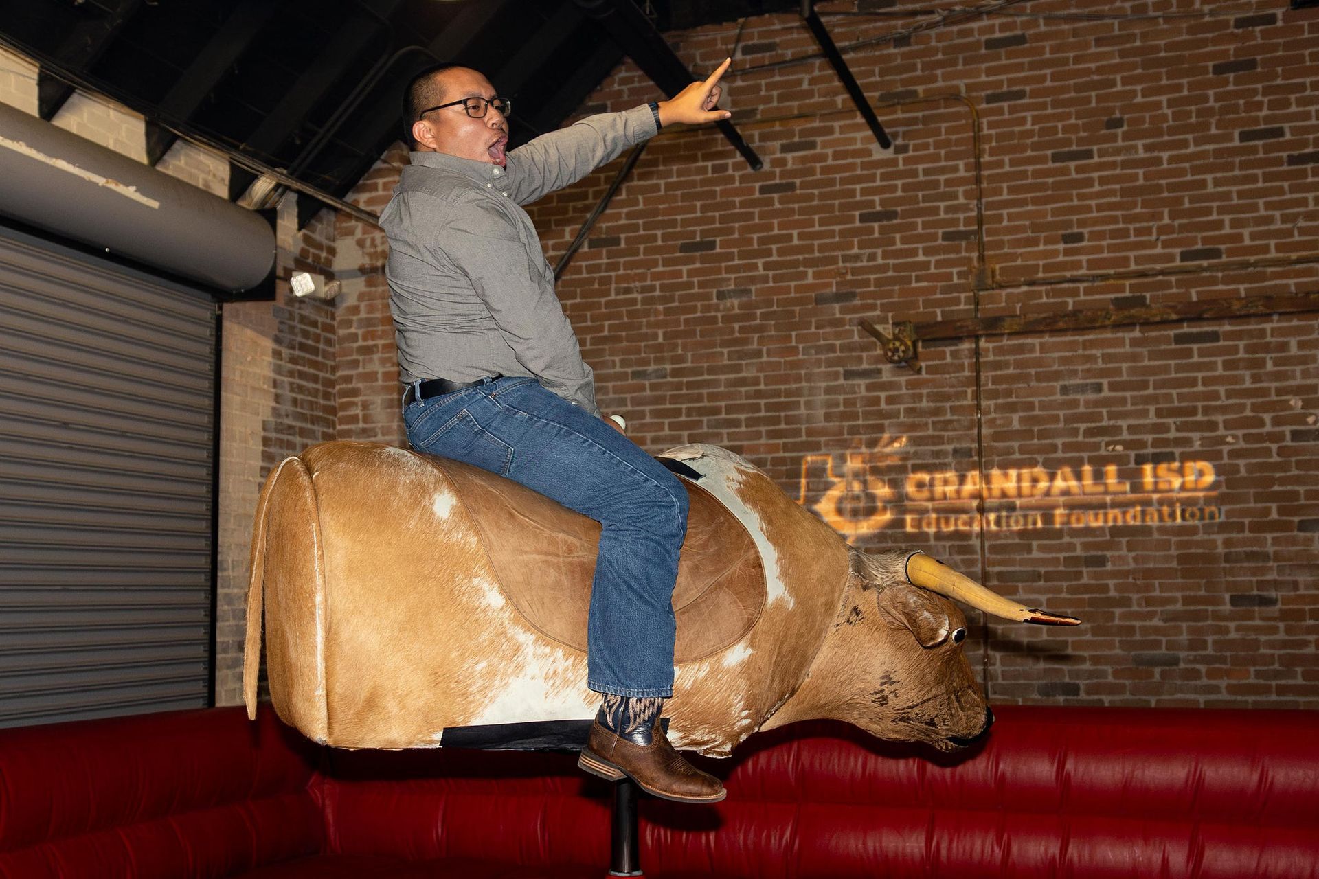 Man riding a mechanical bull indoors, pointing up. Brick wall background, logo visible.
