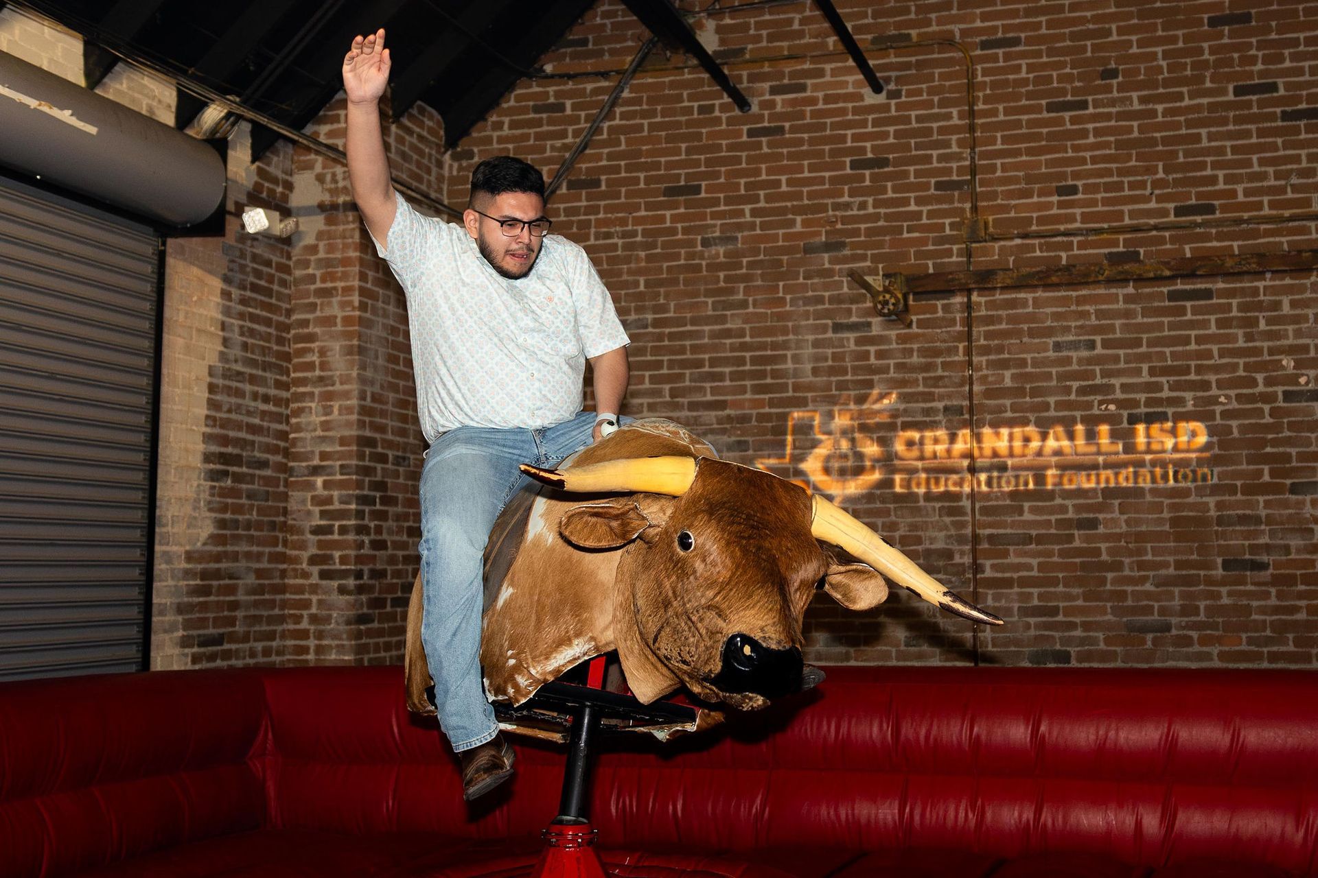 Man riding a mechanical bull, raising his arm with a brick wall background.