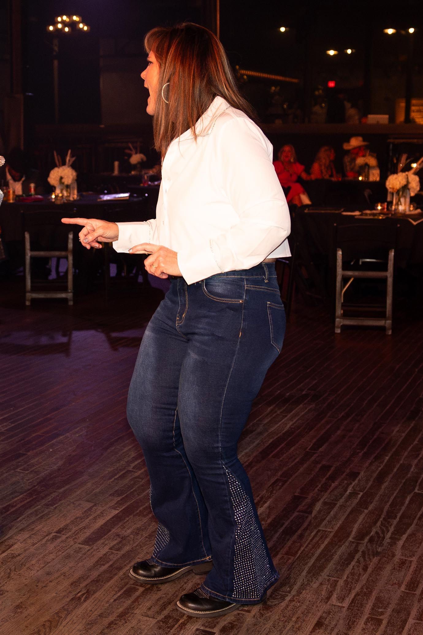 Woman dancing, wearing jeans with embroidered detail and a white shirt, pointing to the left in a dark room.