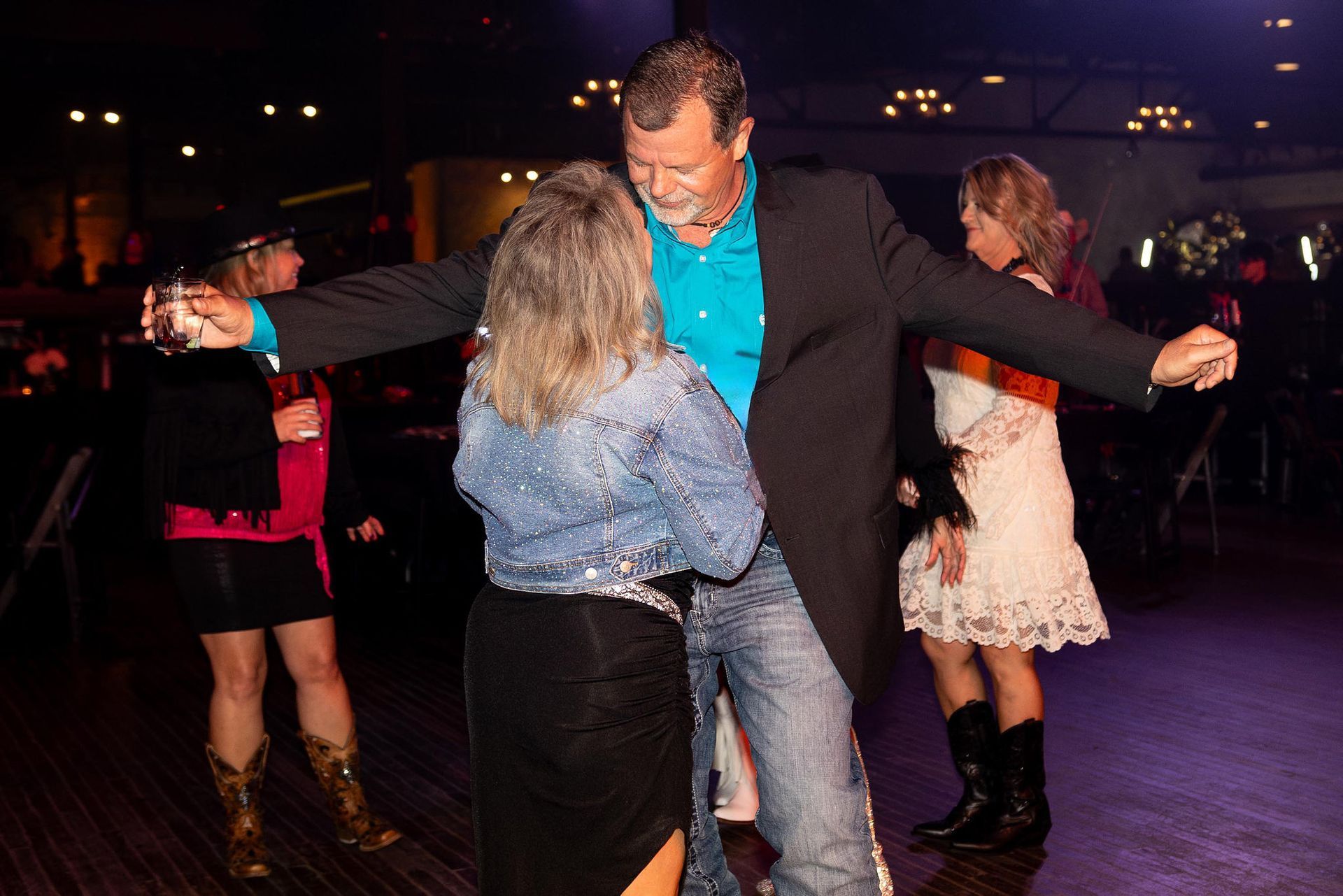 People dancing at an event; a man in a jacket and jeans dances with a woman wearing a denim jacket.