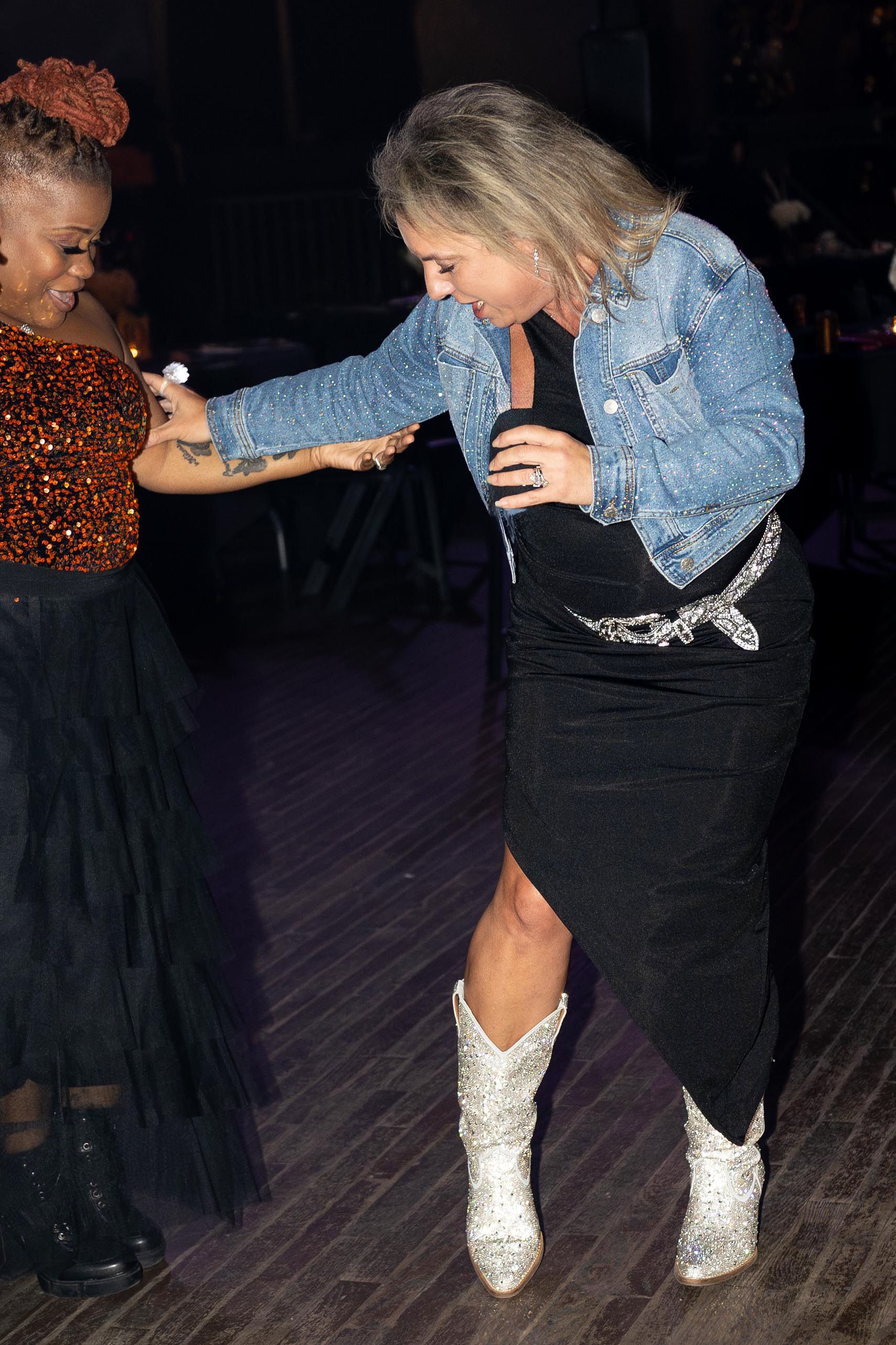 Woman in denim jacket and boots dances with another woman in an orange top and black skirt.
