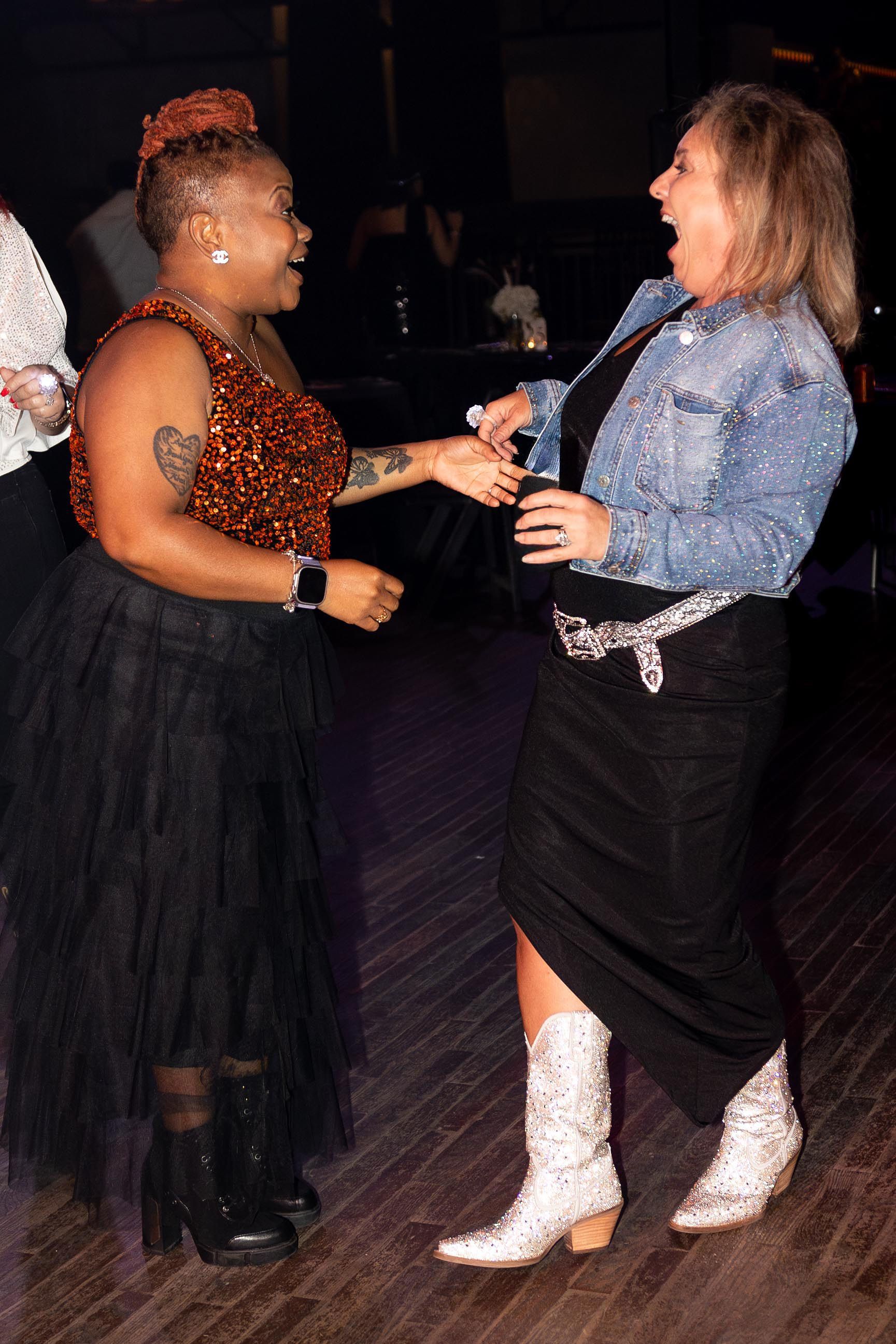 Two women laughing, dancing. One in a sparkly top and tutu, the other in a denim jacket, skirt, and boots.