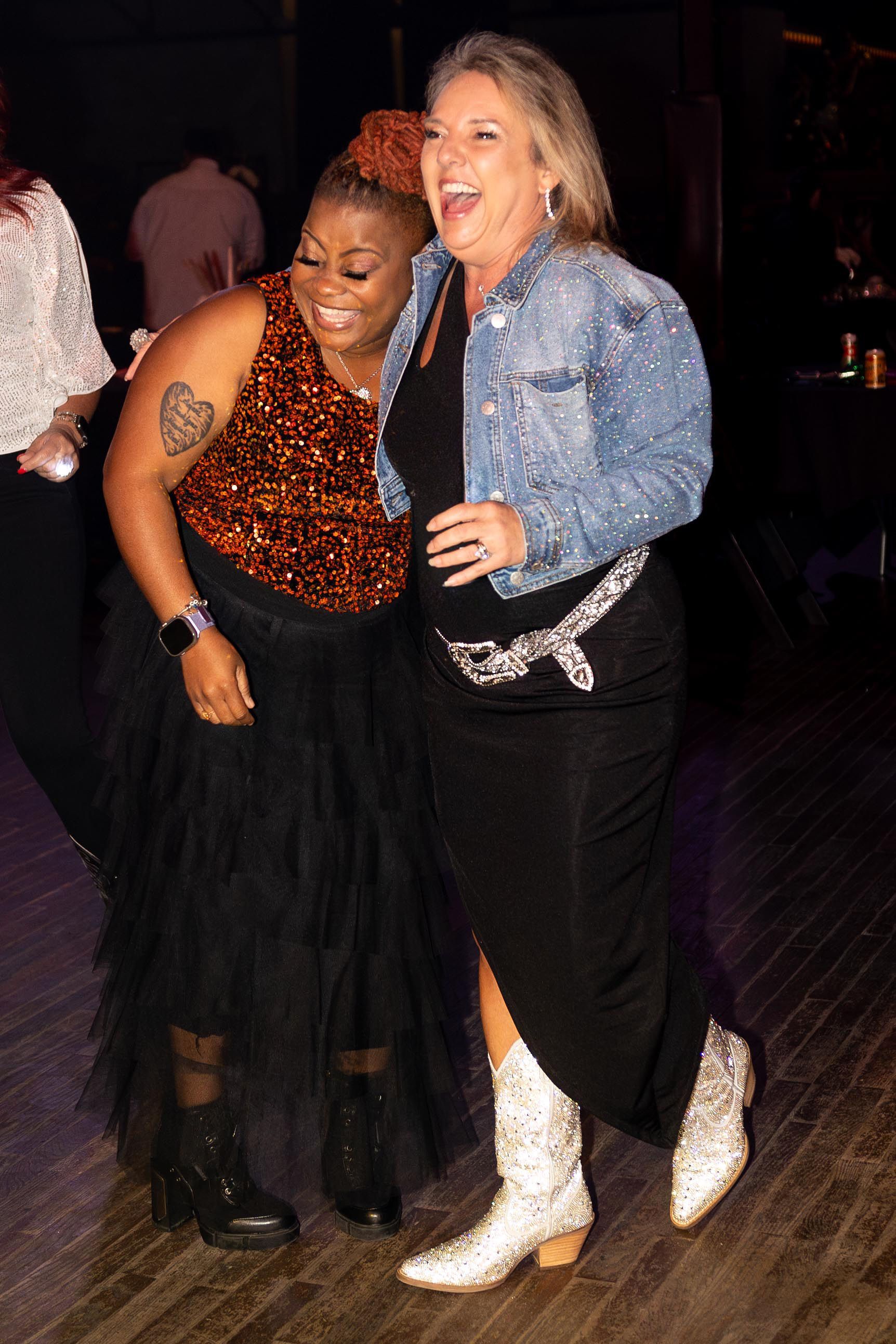 Two women laughing, one in an orange sequin top, the other in a jean jacket, dancing at a party.